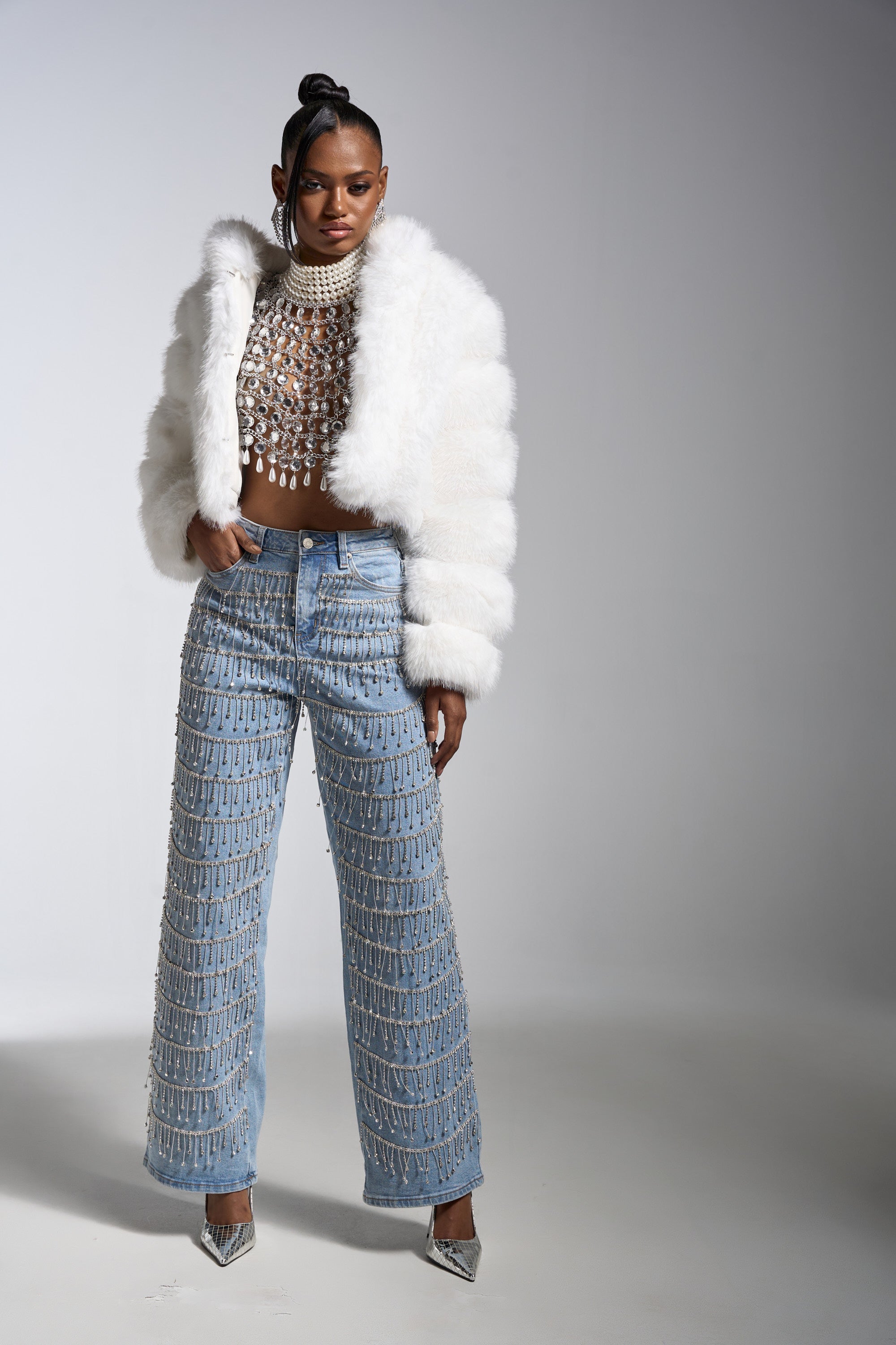 A woman poses in a white faux fur jacket, beaded crop top, DRIP DRIP EMBELLISHED DENIM high-rise rhinestone fringe jeans with silver accents, and pointed silver heels against a plain light gray background.