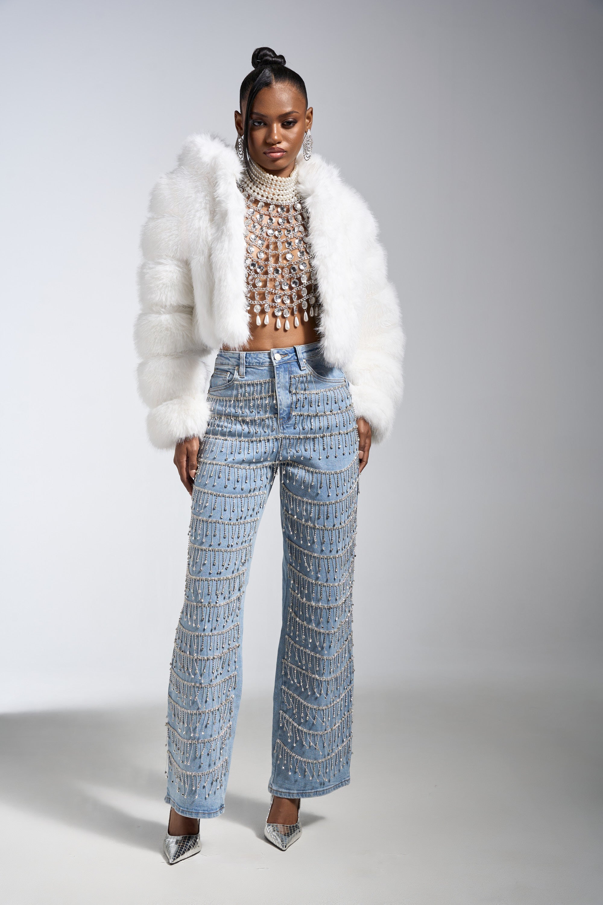 A woman poses confidently in the DRIP DRIP EMBELLISHED DENIM with crystal rhinestone fringe, paired with a fuzzy white jacket, jeweled crop top, and silver pointed heels against a plain, light background.