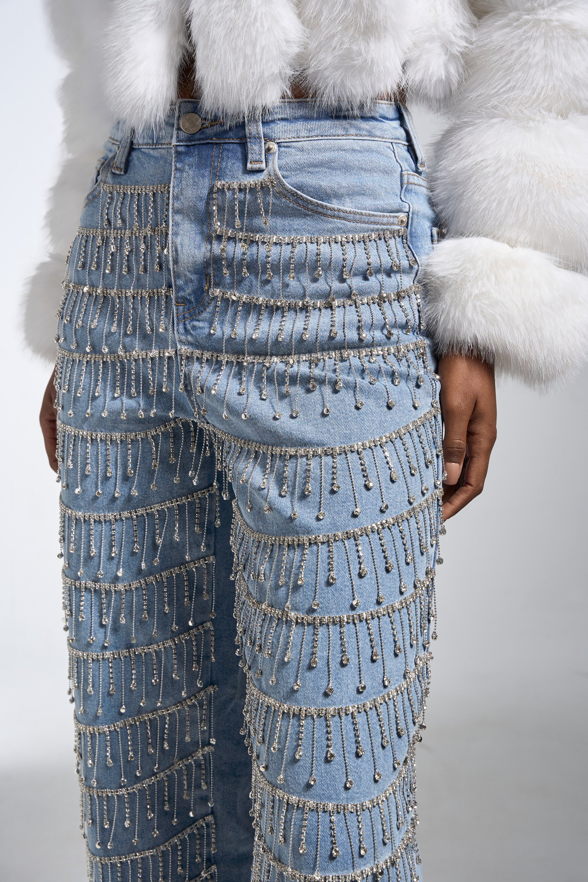 A person wears the DRIP DRIP EMBELLISHED DENIM—high-rise straight jeans adorned with silver beads and chains—paired with a white faux fur jacket. The image, cropped at the waist and mid-thigh, highlights the outfit's intricate details.
