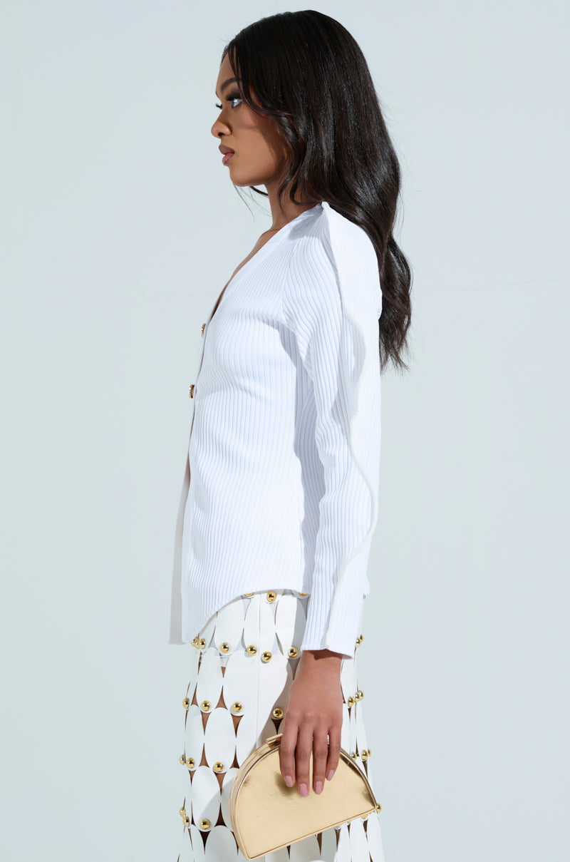 A woman with long, dark hair stands in profile against a light background, wearing a white blouse and patterned skirt. She holds a small gold purse, embodying elegance akin to the sophistication of the WAVY CARDIGAN IN WHITE's ribbed knit fabrication.