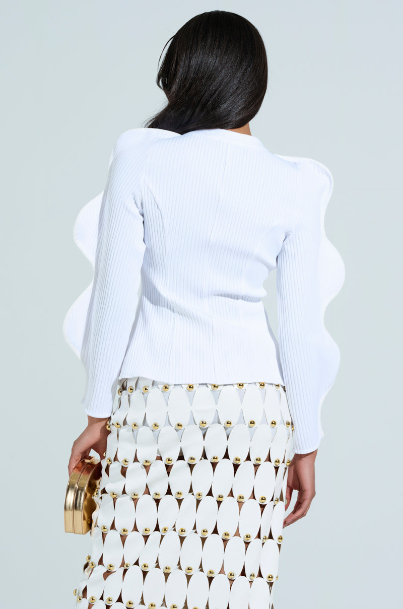 A person with long dark hair wears the WAVY CARDIGAN IN WHITE, expertly crafted from ribbed knit, paired with a white skirt featuring gold accents. They hold a small gold clutch and face away from the camera against a light gray backdrop.