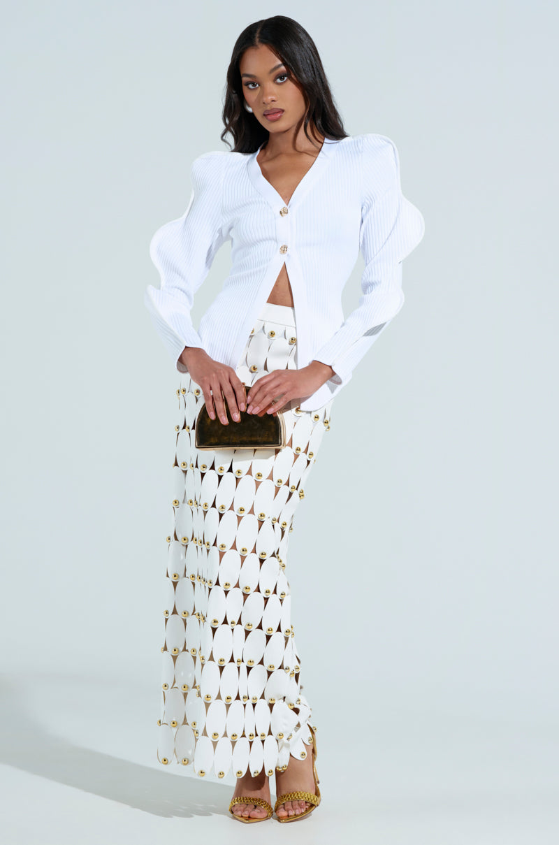 A woman poses against a neutral background in a stylish ensemble: the WAVY CARDIGAN IN WHITE with structured shoulders, paired with a long geometric-patterned skirt. She holds a small purse gracefully and stands confidently in gold heels.