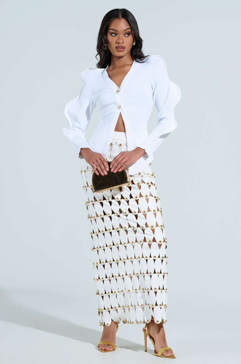 Against a plain backdrop, a woman wears a white blouse with puffed sleeves, a gold and white geometric skirt, gold heels, and holds a brown clutch. Her outfit is complemented by the WAVY CARDIGAN IN WHITE made from ribbed knit fabric.