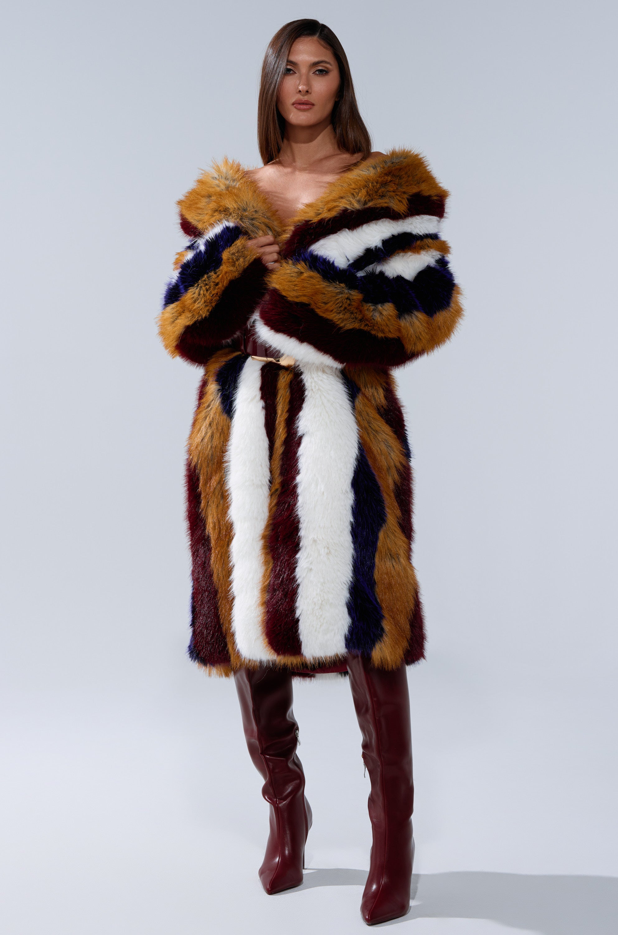 A woman stands confidently in the ALAIA STRIPED FAUX FUR coat with brown, white, navy, and gold stripes, paired with knee-high maroon boots against a plain light gray background.