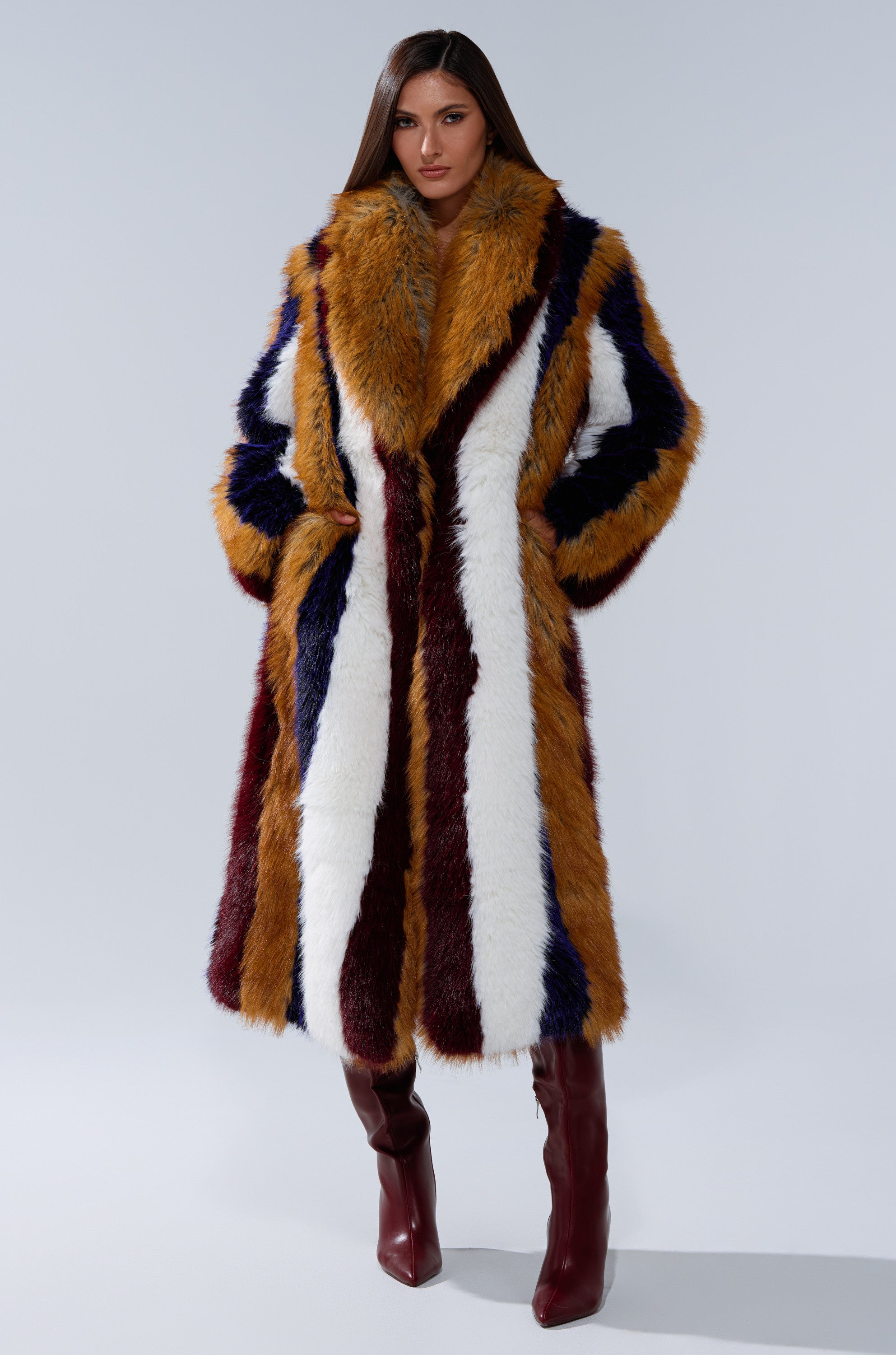 A woman stands confidently in the ALAIA STRIPED FAUX FUR longline coat with bold vertical stripes of mustard yellow, burgundy, navy blue, and white, paired with tall burgundy boots and straight brown hair.