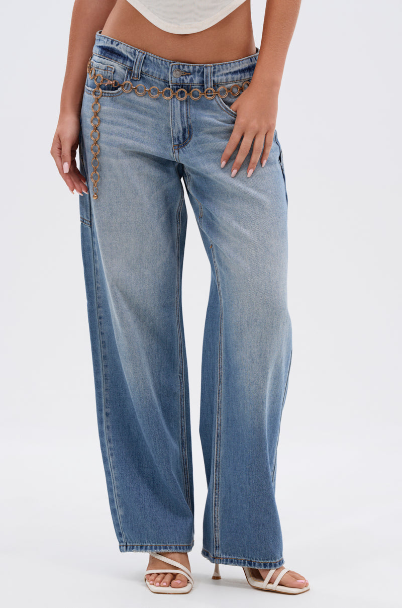 Someone stands against a plain light background wearing the RELAX WITH ME WIDE LEG DENIM jeans with a brown grommet belt, a partially visible white top, and white heeled sandals.