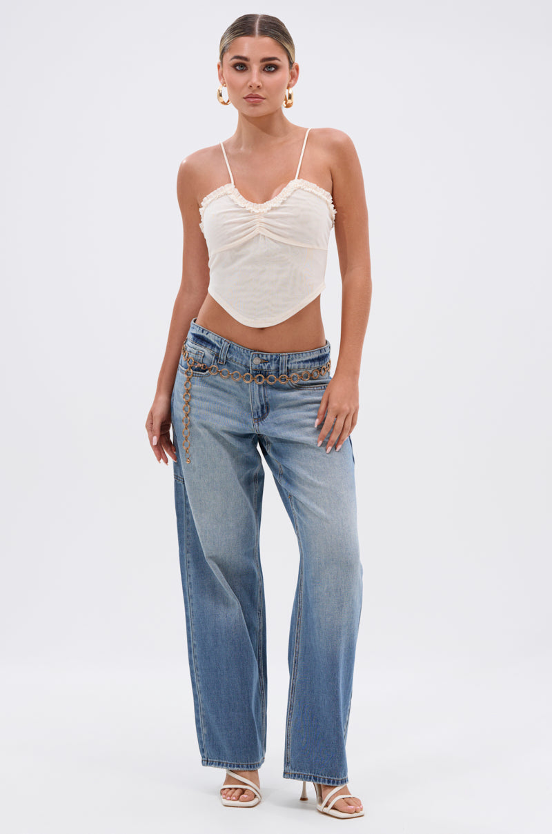 A woman poses confidently against a plain background, wearing the RELAX WITH ME WIDE LEG DENIM, a cream ruched camisole, chain belt, white heeled sandals, large hoop earrings, and her hair slicked back.