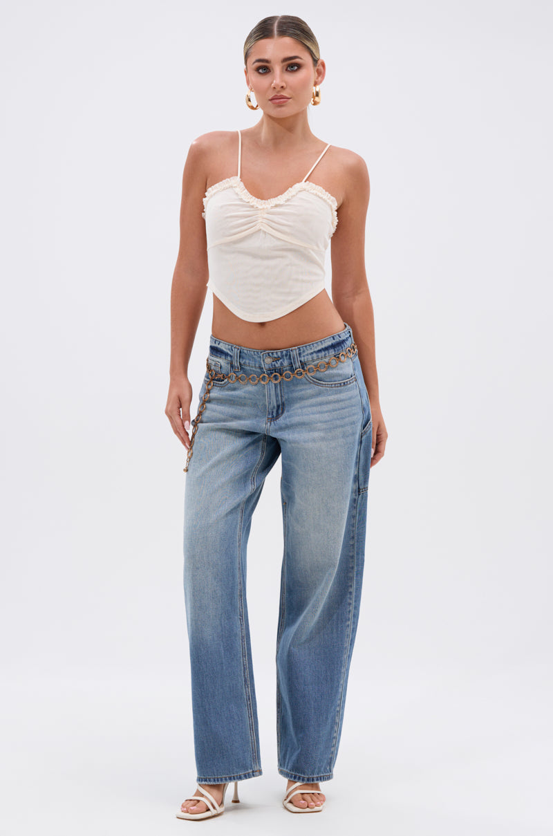 A woman stands against a white background wearing a cream cropped camisole, RELAX WITH ME WIDE LEG DENIM jeans with a chain belt, white heeled sandals, and gold hoop earrings.