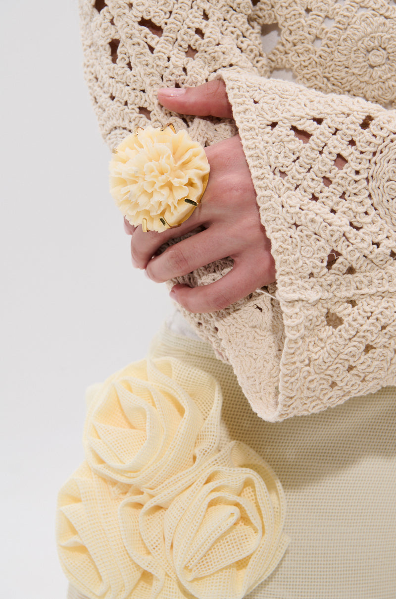 A person wearing a cream crocheted top with flared sleeves holds their hands together, showcasing the IN BLOOM RING. They pair it with a skirt adorned with fabric roses, all in soft pale yellow tones for a coordinated look.