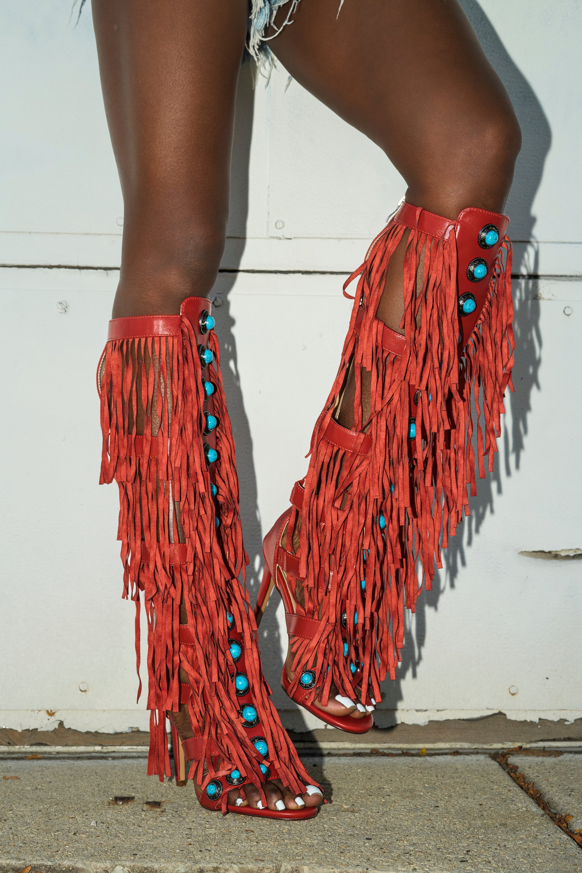 A person wears the AZALEA WANG AMADEO RED SANDAL—knee-high, gladiator-inspired boots with dramatic fringe and turquoise details—styled with cut-off denim shorts for a bold, modern look against a white wall.