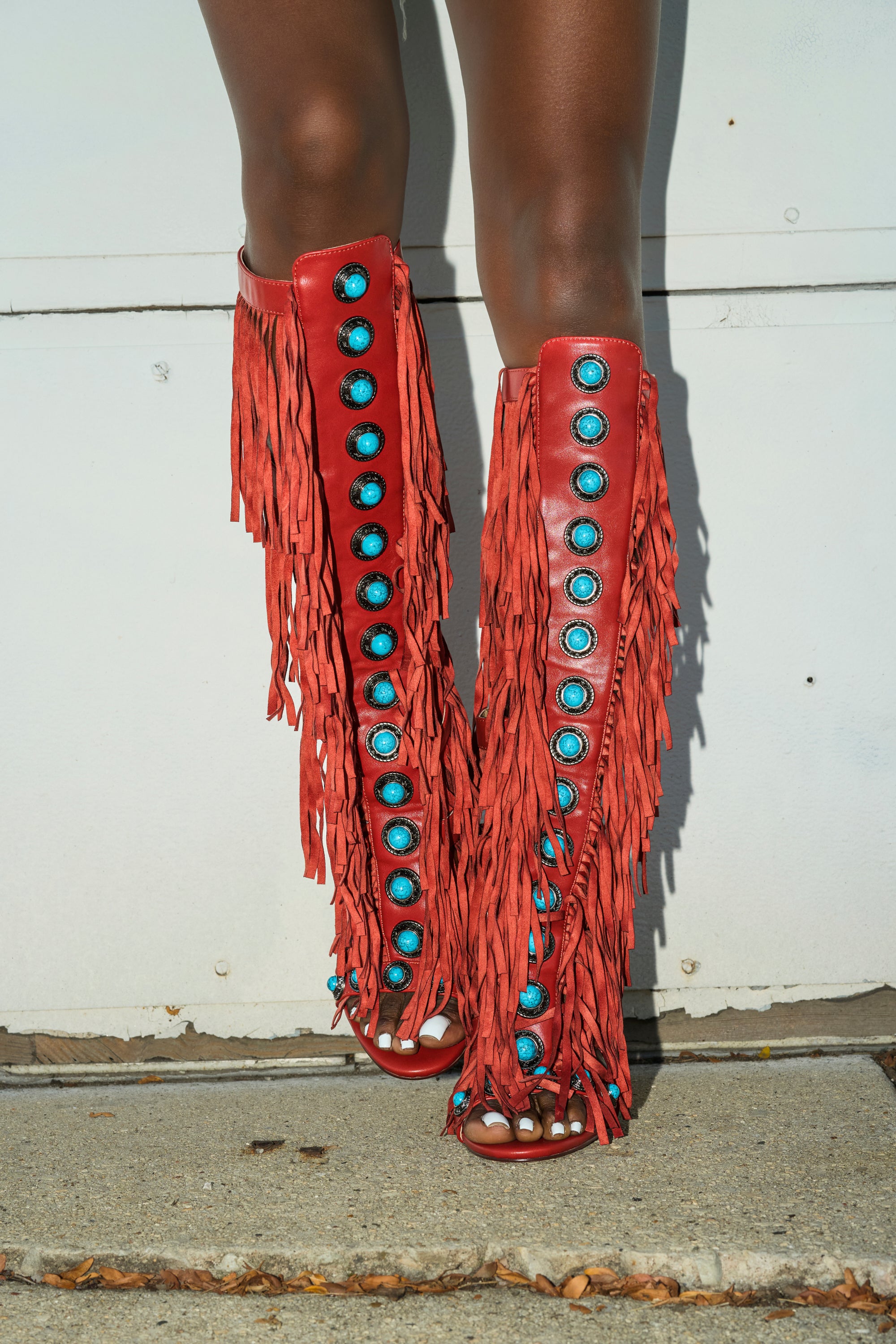 A person stands on a concrete sidewalk in front of a white wall, wearing the AZALEA WANG AMADEO RED SANDAL. Only their legs and feet are visible.