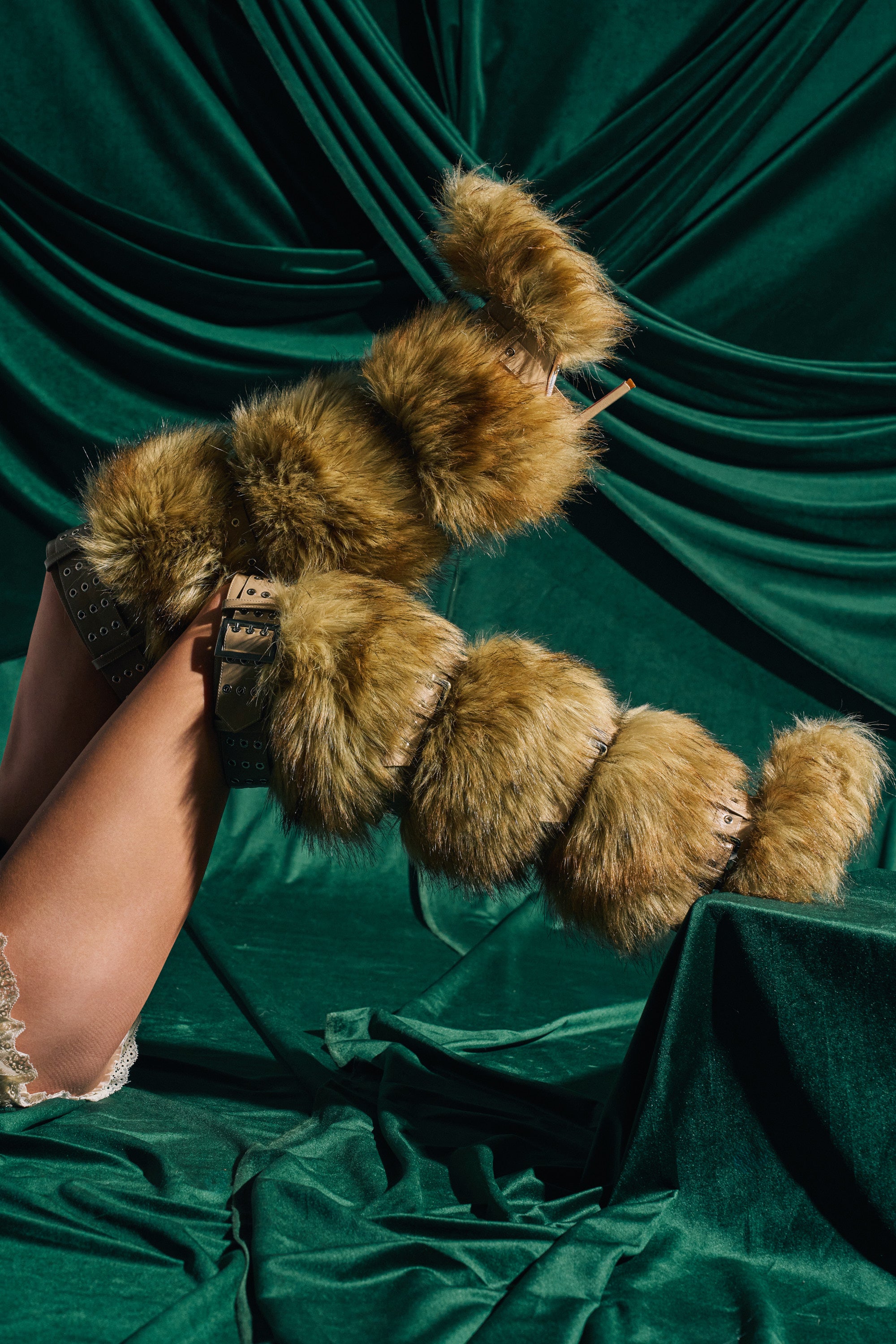 A pair of legs in AZALEA WANG AMALBERT BROWN MULTI FUR AND BUCKLES BOOTS, featuring large fluffy brown faux fur bands, are raised against draped dark green velvet fabric.