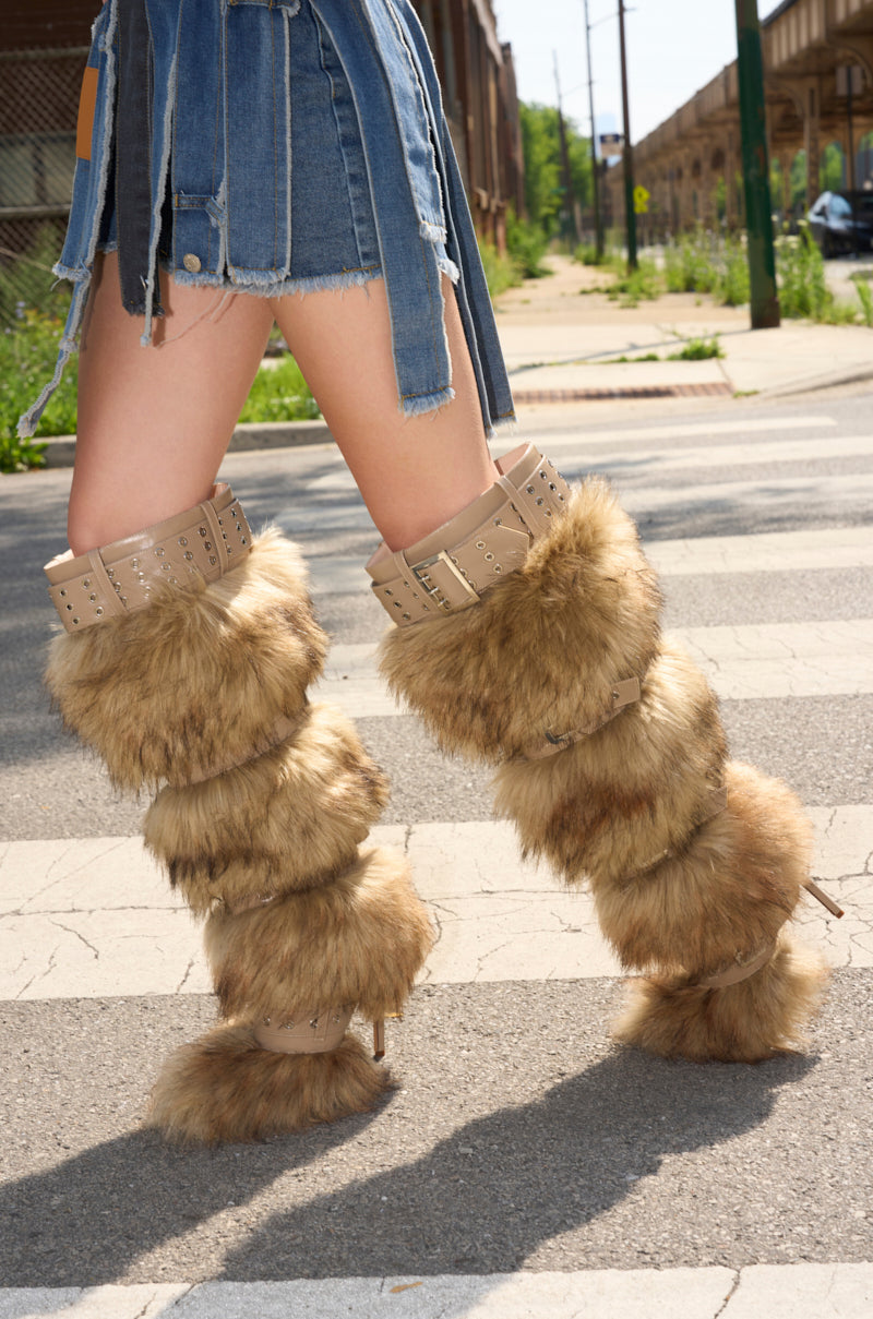 A person wearing a short, frayed denim skirt and AZALEA WANG AMALBERT BROWN MULTI FUR AND BUCKLES BOOT stands on a crosswalk outdoors, with urban buildings and greenery visible in the background.