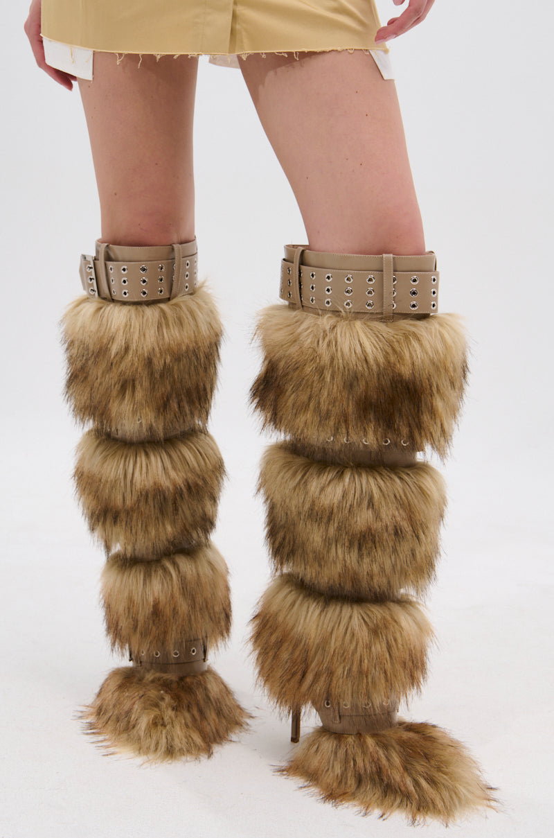 A person stands on a light surface wearing tan shorts and the AZALEA WANG AMALBERT BROWN MULTI FUR AND BUCKLES BOOT—knee-high, faux fur boots with buckled straps.