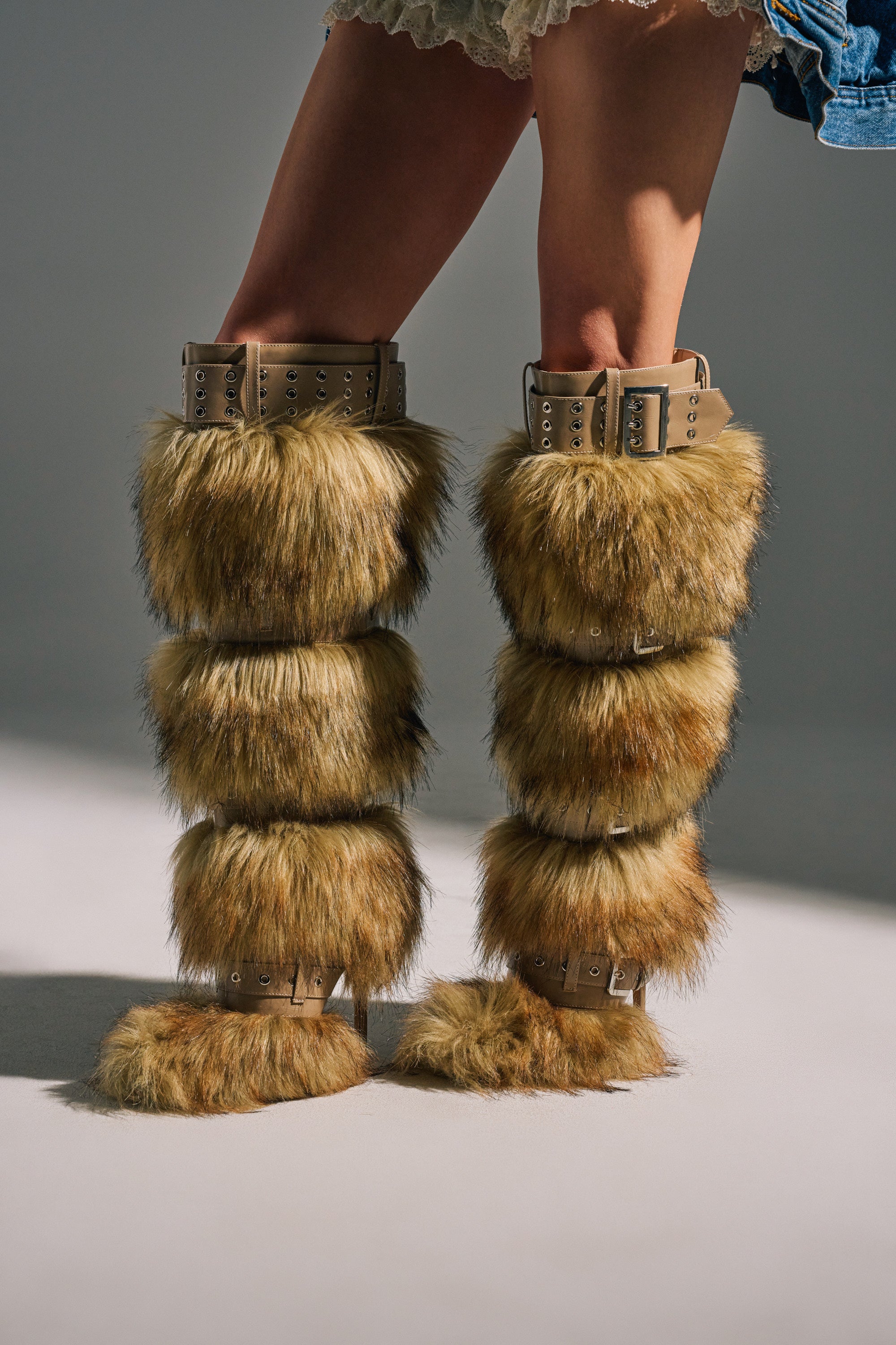 The lower legs and part of a denim skirt are shown as a person wears the AZALEA WANG AMALBERT BROWN MULTI FUR AND BUCKLES BOOT, featuring tall, tan faux fur with multiple sections and buckle details on a neutral background.