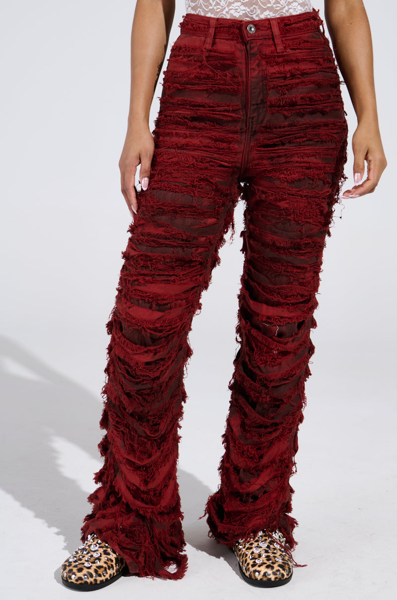 Someone in shredded ON FIRE DISTRESSED DENIM PANT, a red mid-rise design with frayed texture, pairs it with leopard print slip-ons against a plain backdrop. The high-waisted pants offer a stylish look, while the top is partially visible.
