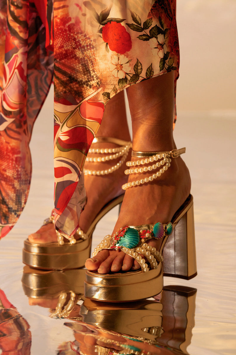 A close-up shows someone wearing AZALEA WANG ARLETH GOLD SANDALS with chunky heels adorned with pearls and turquoise beads. The ankle straps feature pearl details, perfectly complementing a flowing floral and abstract patterned dress in the background.