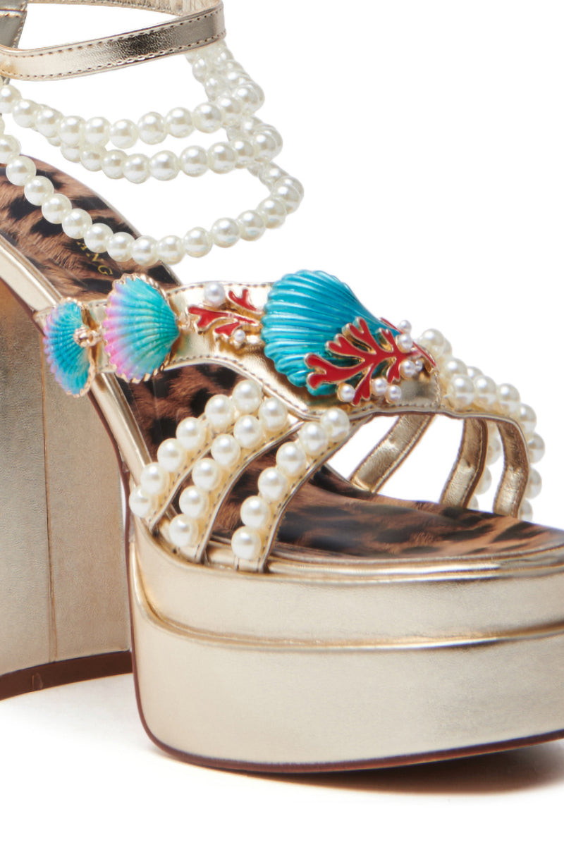 A close-up of the AZALEA WANG ARLETH GOLD SANDAL, featuring a chunky heel and a platform with pearl beads. The straps glisten with colorful seashell embellishments, while the lining displays an animal print pattern.