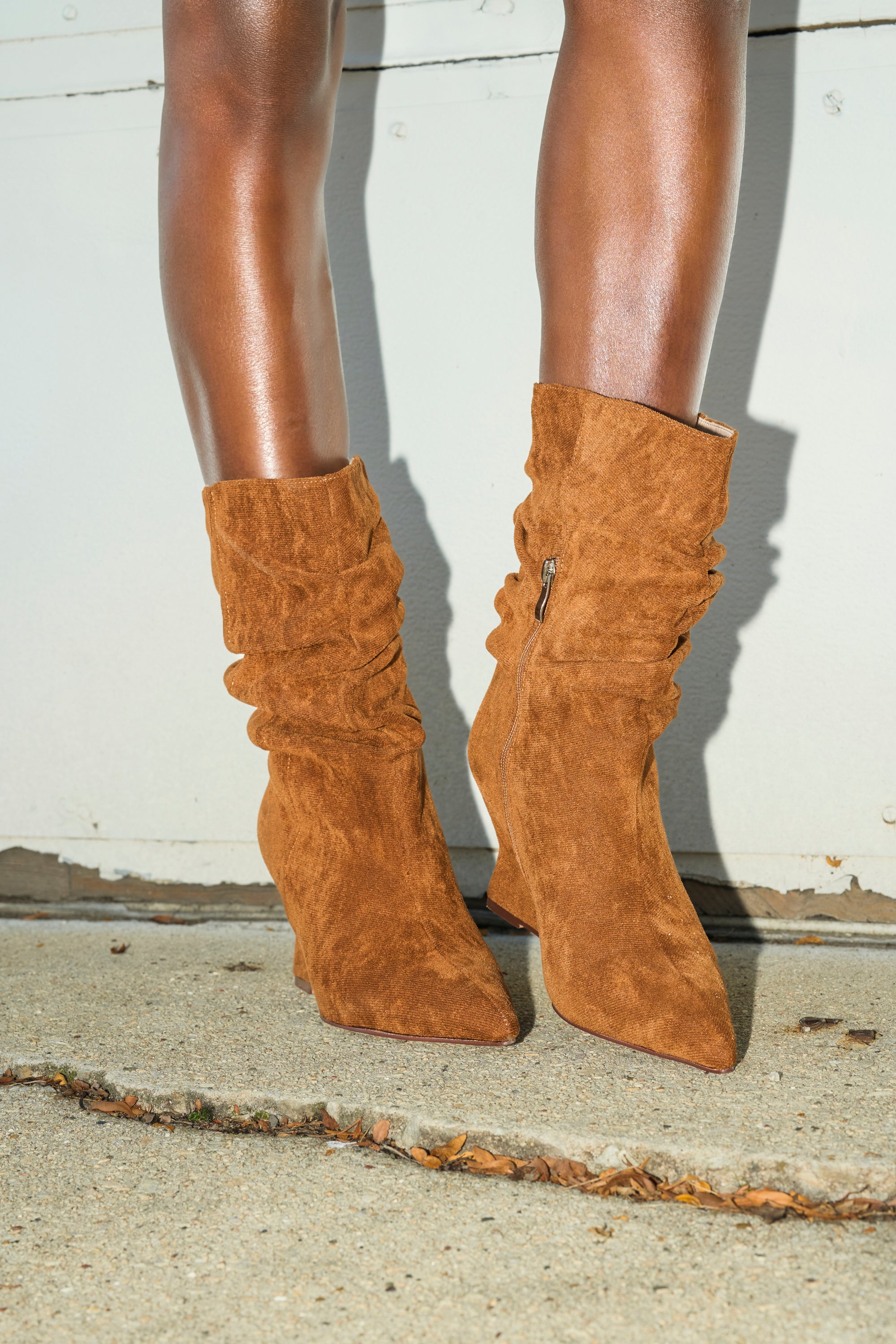 Close-up of a person wearing the AZALEA WANG ASAF Ruched Wedge Bootie in tan, featuring a pointed toe and slouchy suede style, standing on concrete near a white wall.