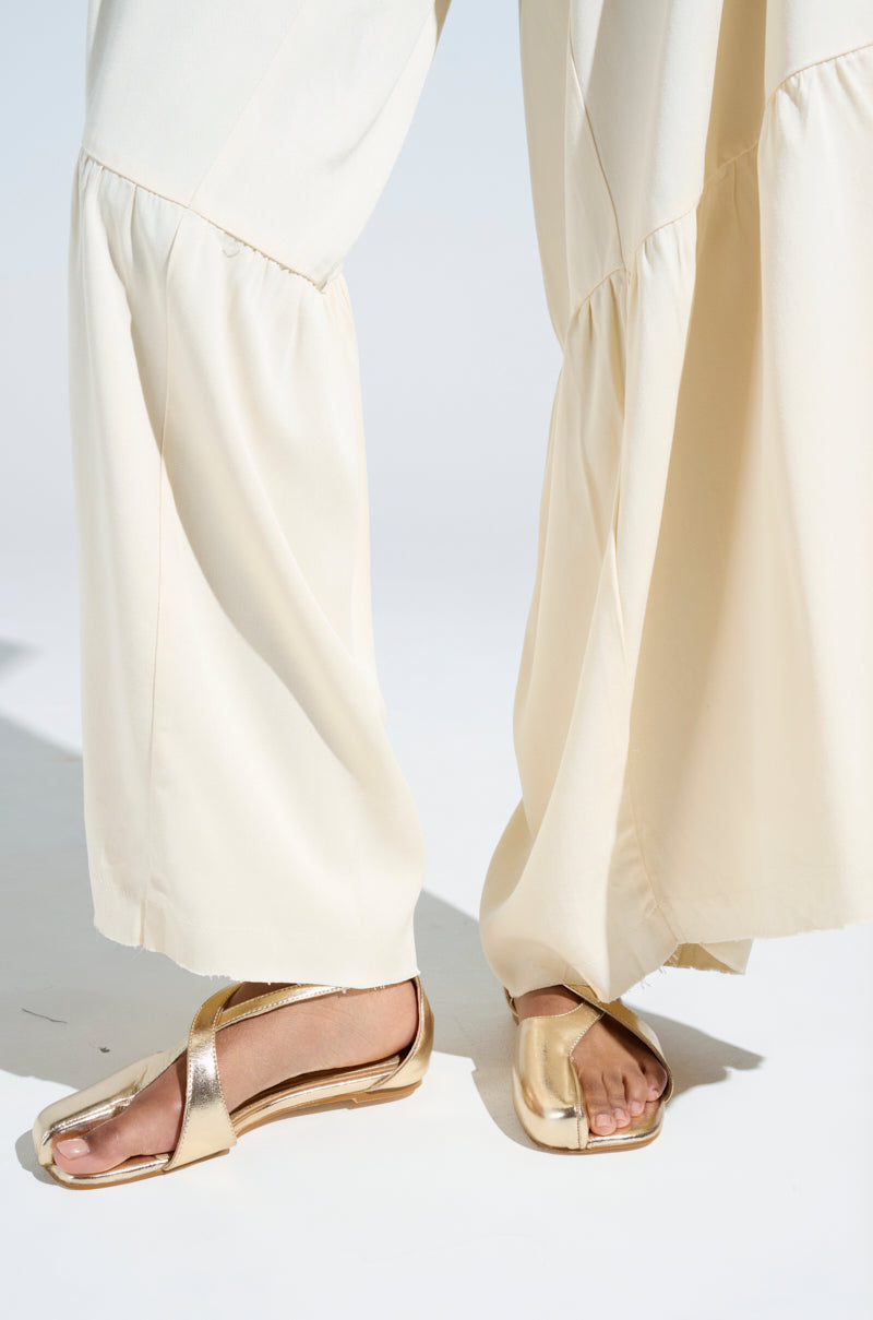 The image features a close-up of a person wearing cream-colored, flowy pants and the AKIRA AULORA Gold Flat Sandals with crisscross straps and faux leather, emphasizing their casual yet elegant style. The neutral background keeps the focus on the stylish footwear.
