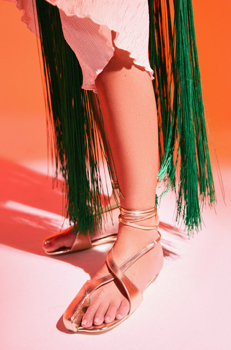 A person wearing the AKIRA AULORA Gold Flat Sandal with peep toe and wrap-around ankle straps stands on a pink and orange surface, styled with a white pleated skirt and long green fringe.