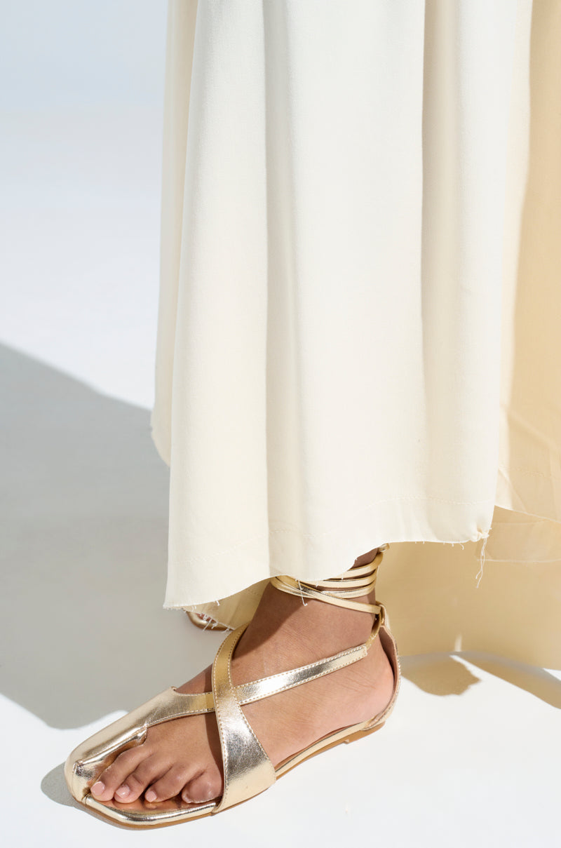 A person stands on a light background, showcasing the AKIRA AULORA gold flat sandals with peep toe and crisscross straps. Their lower legs are visible, dressed in a flowing cream-colored fabric.