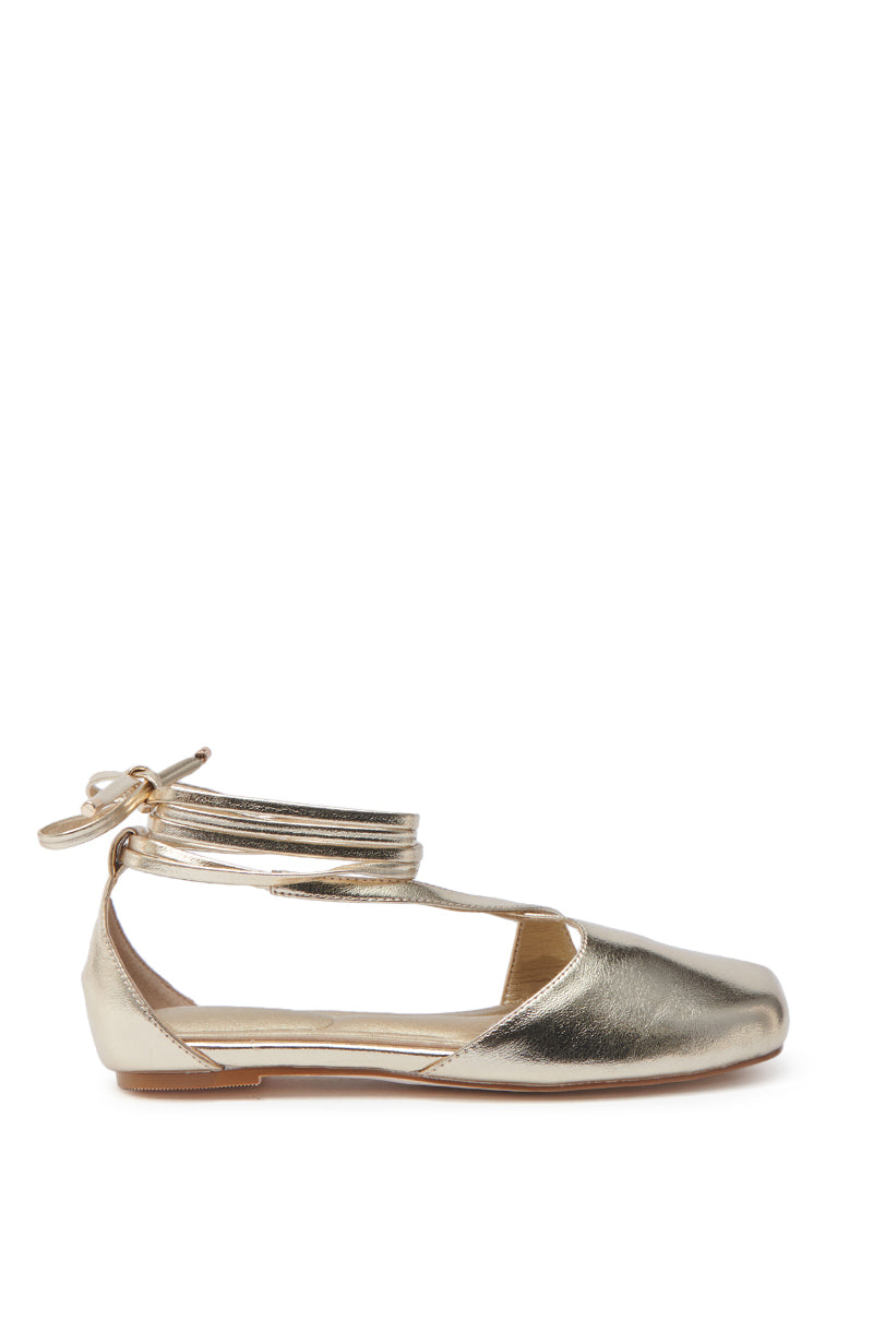 Side view of the AKIRA AULORA Gold Flat Sandal with peep toe featuring crisscross straps and ankle wraparound ties in a bow. The faux metallic gold leather shines, complemented by its flat sole.