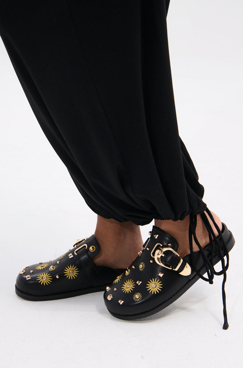 Wearing black pants with drawstring cuffs, a person showcases the AZALEA WANG TROUVER BLACK GOLD STARBURST FLAT embellished clogs on a light gray floor.