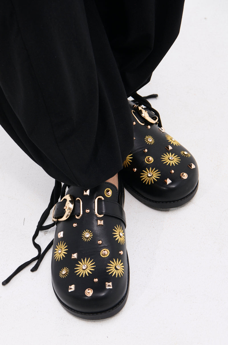 The AZALEA WANG TROUVER BLACK GOLD STARBURST FLAT features faux leather with gold starbursts, studs, and buckles. These statement clogs pair well with loose black pants and showcase eye-catching decorative metal accents.