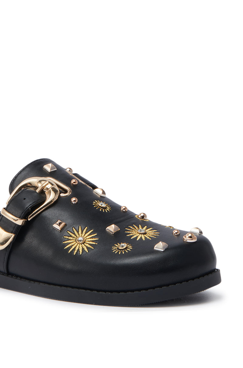 A close-up of the AZALEA WANG TROUVER BLACK GOLD STARBURST FLAT shows its faux leather, gold buckles, decorative studs, and starburst designs. This flat-soled clog features multiple gold accents on a sleek black surface.