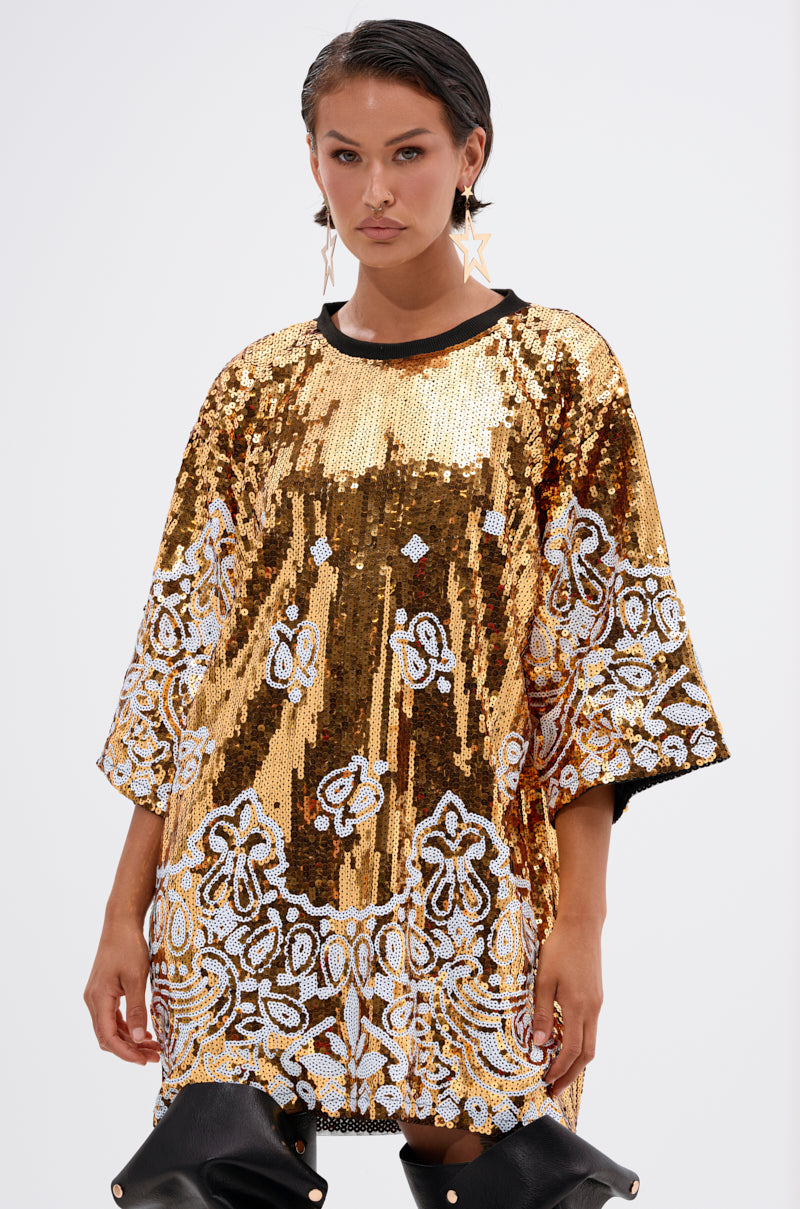 A person wears the BOUJEE BANDANA SEQUIN MINI DRESS, featuring gold embellishments and white floral patterns, paired with large star-shaped earrings and black pants, standing against a plain light background.