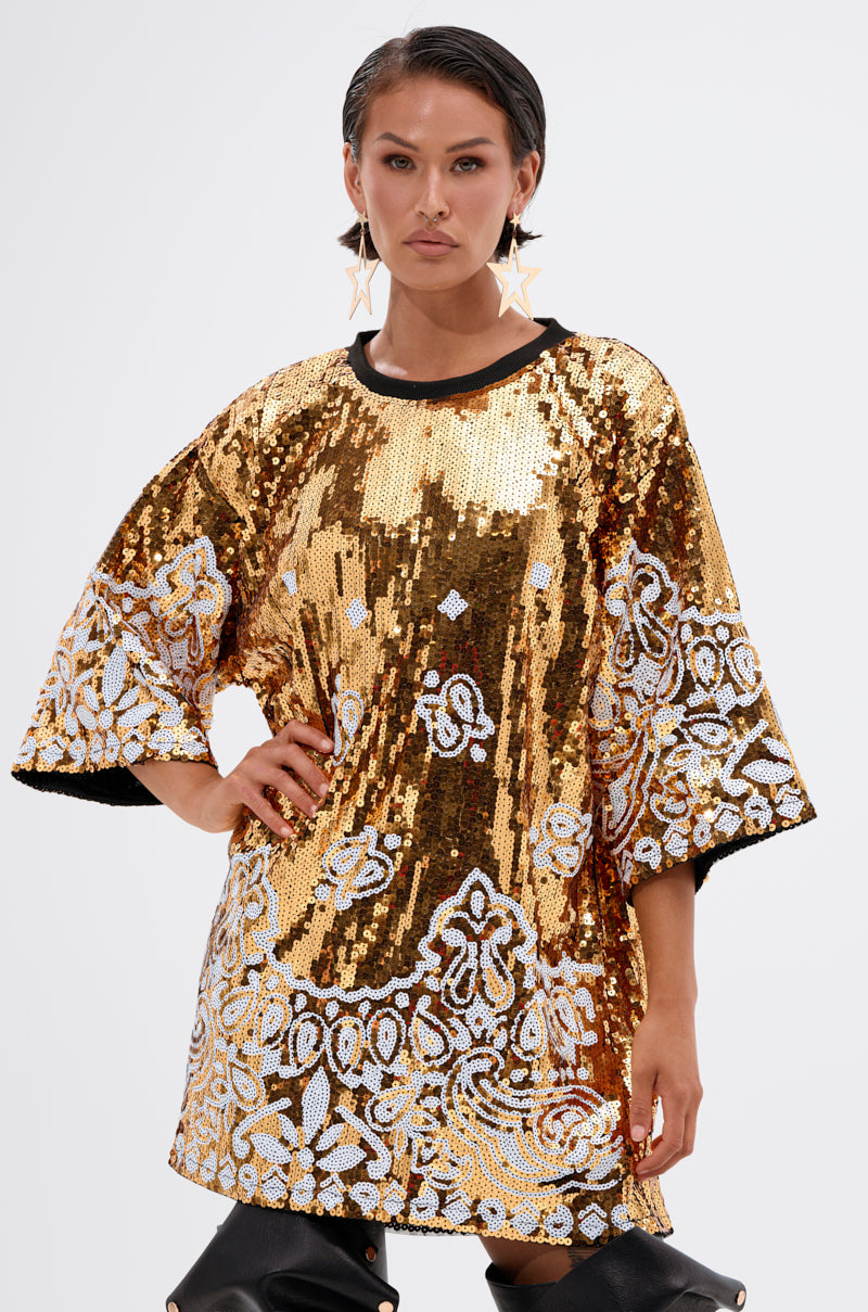 A woman poses confidently in the BOUJEE BANDANA SEQUIN MINI DRESS with wide sleeves and black trim, styled with star-shaped earrings and black high boots, set against a plain light background.