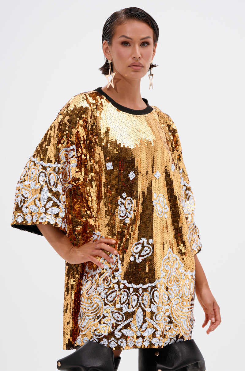 A person with short dark hair wears the BOUJEE BANDANA SEQUIN MINI DRESS, featuring gold embellishments and decorative patterns, paired with star-shaped earrings, posing confidently against a light background.