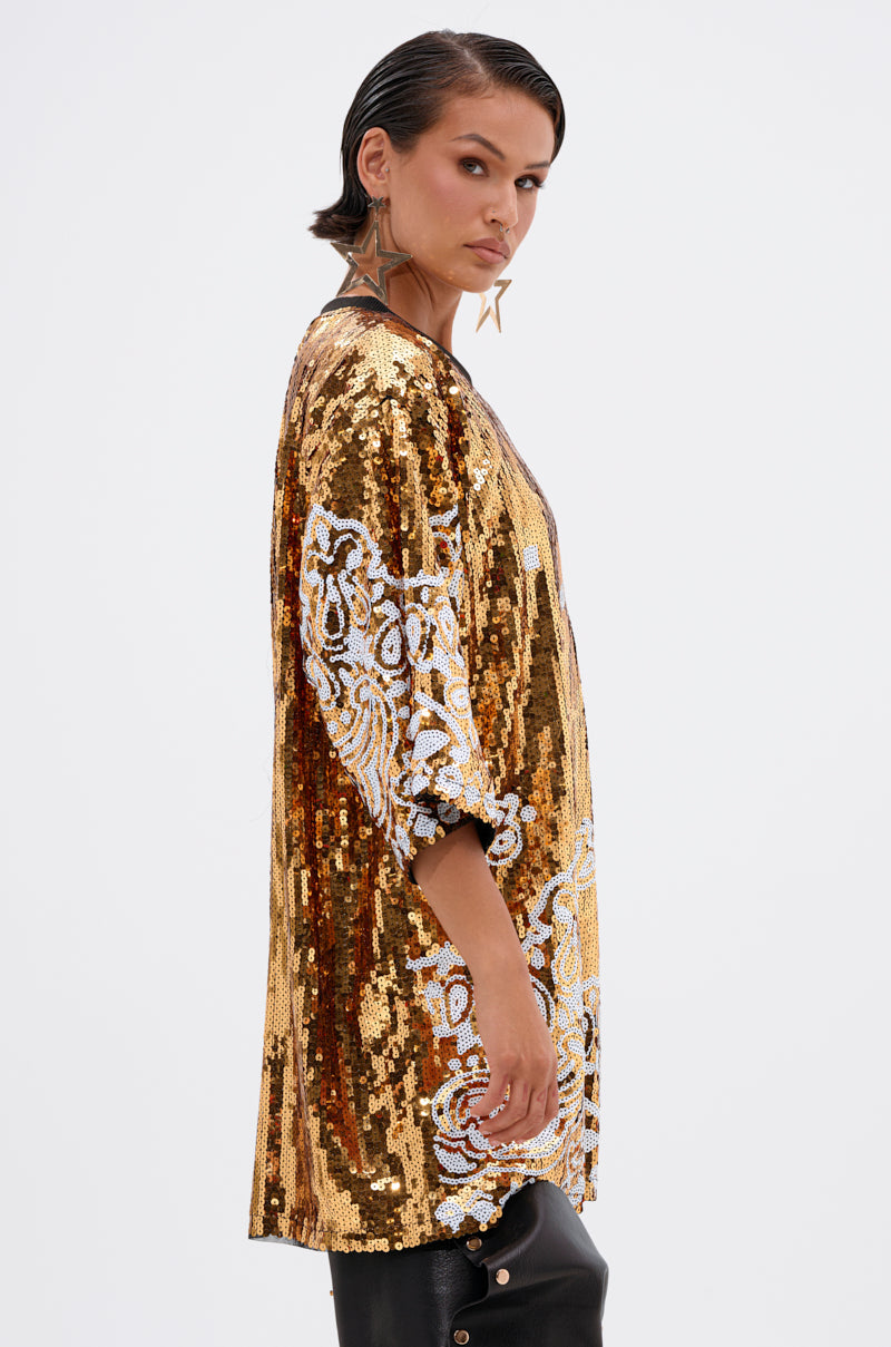 A person with short dark hair wears the BOUJEE BANDANA SEQUIN MINI DRESS with gold embellishments and white sequin floral patterns, paired with black earrings and pants, standing sideways and looking at the camera against a white background.