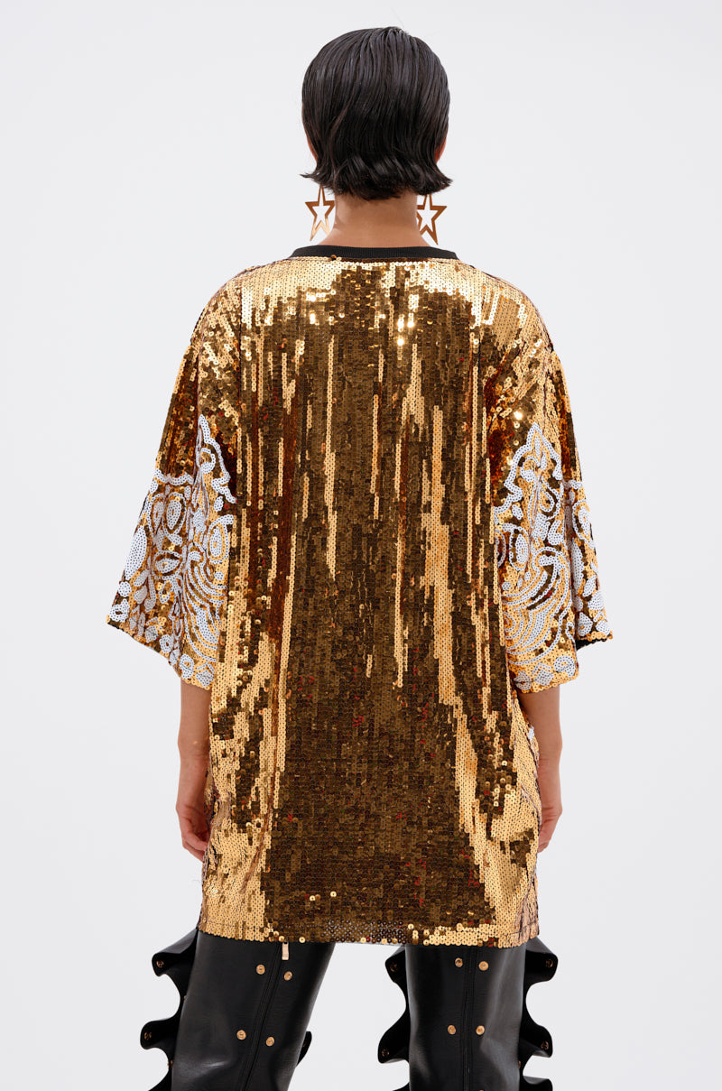 A person with short dark hair and star-shaped earrings stands against a plain white background, wearing the BOUJEE BANDANA SEQUIN MINI DRESS—loose, shiny, gold-embellished with white lace sleeves—paired with black ruffled pants.