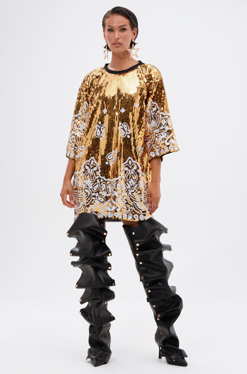 A person stands facing forward, wearing the BOUJEE BANDANA SEQUIN MINI DRESS—a gold embellished dress with white and black floral patterns—paired with oversized black slouchy thigh-high boots, slicked-back hair, and star-shaped earrings.