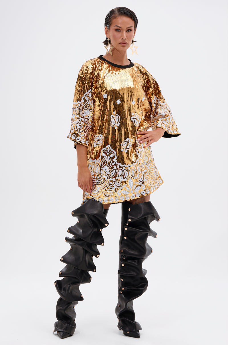 A woman poses confidently in the BOUJEE BANDANA SEQUIN MINI DRESS, a gold embellished piece with white floral patterns, paired with statement ruffled thigh-high boots and large star-shaped earrings, her hair slicked back.