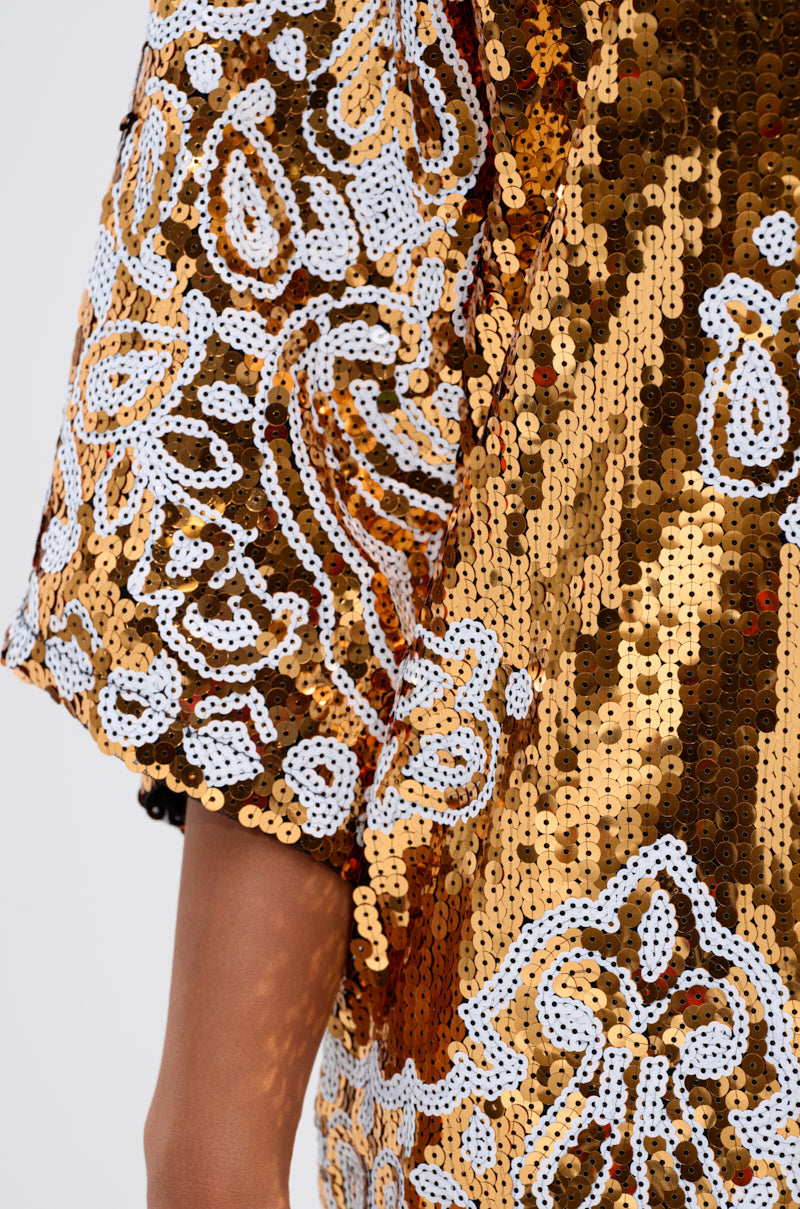 Close-up of a person's arm in the BOUJEE BANDANA SEQUIN MINI DRESS, featuring an ornate gold-embellished sleeve with intricate white bead and sequin patterns. Only part of the arm and shimmering mini dress is visible.