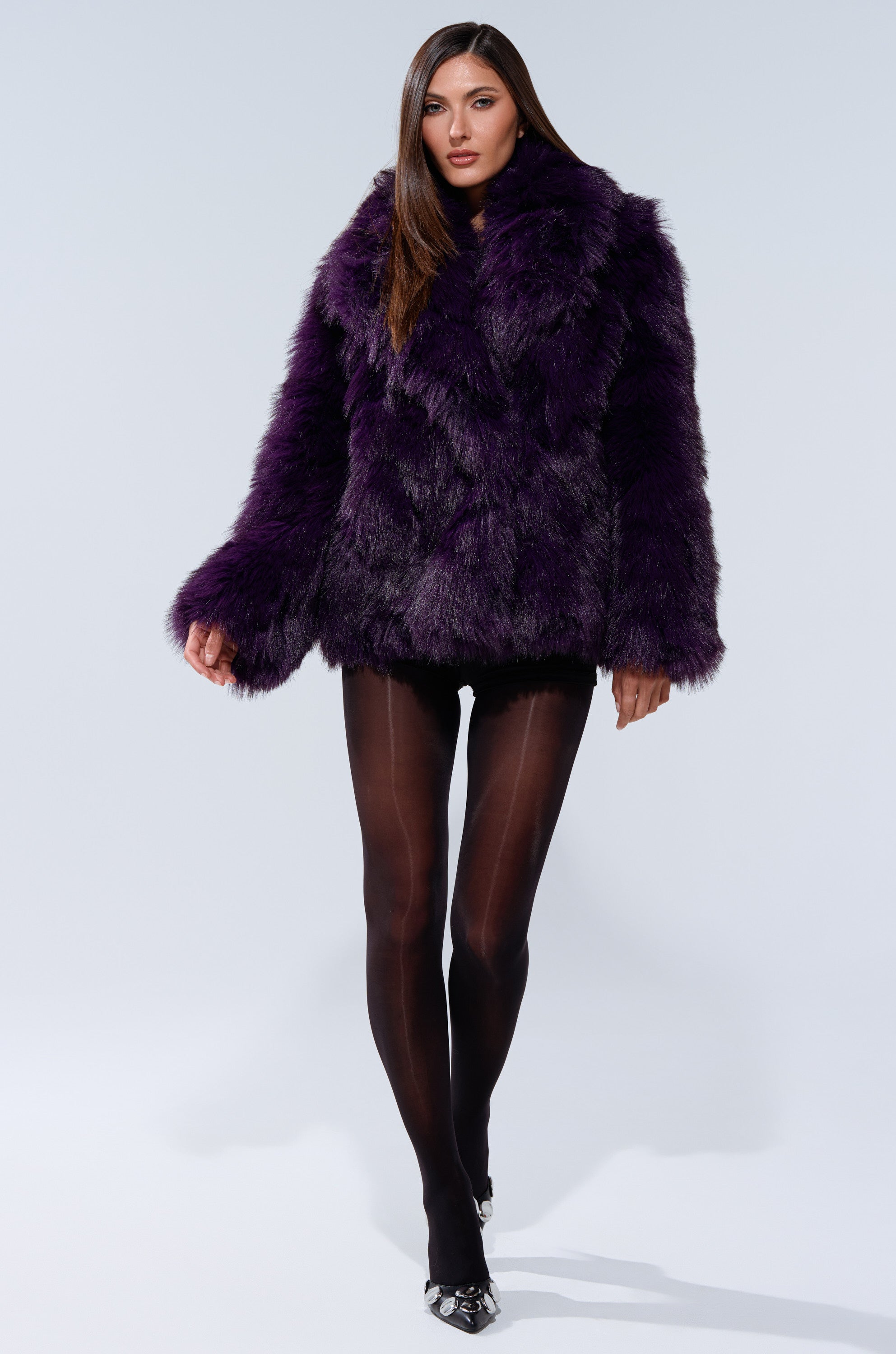 A woman walks confidently against a plain light background wearing the SHORTIE FUR COAT, paired with sheer black tights and pointed shoes.