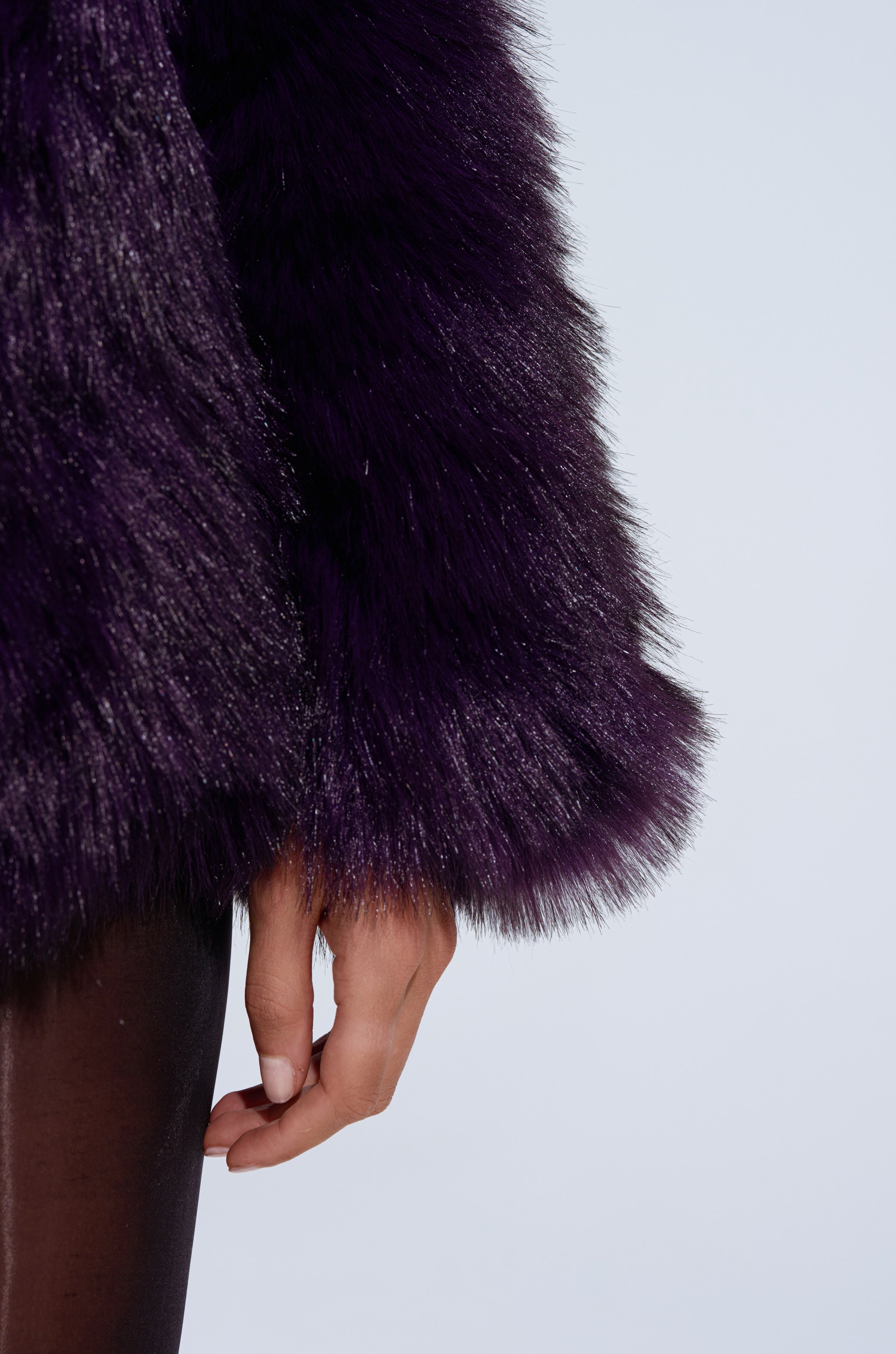 Someone wearing the SHORTIE FUR COAT, an oversized purple faux fur piece, pairs it with shiny black pants and stands with their hand relaxed by their side against a plain light background.