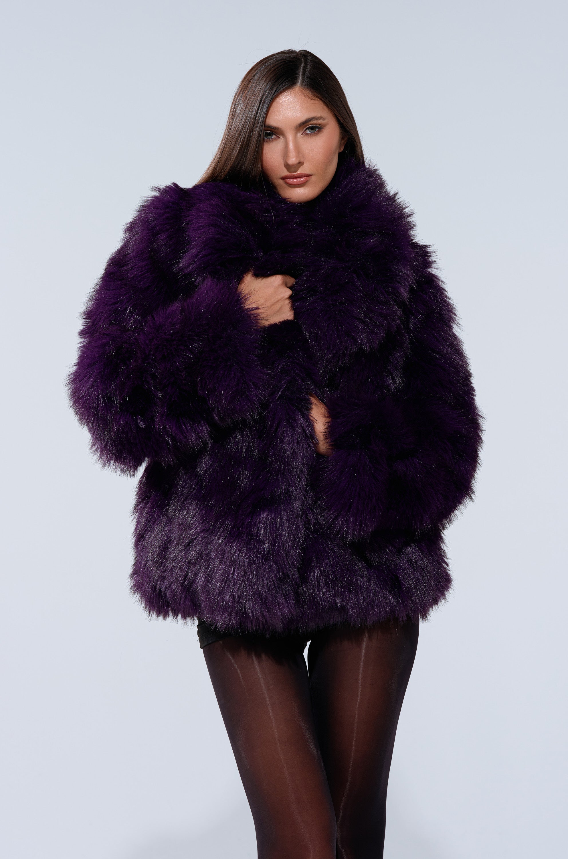 A woman with straight brown hair showcases the SHORTIE FUR COAT—an oversized, luxurious purple faux fur jacket—paired with sheer black tights against a plain light background.