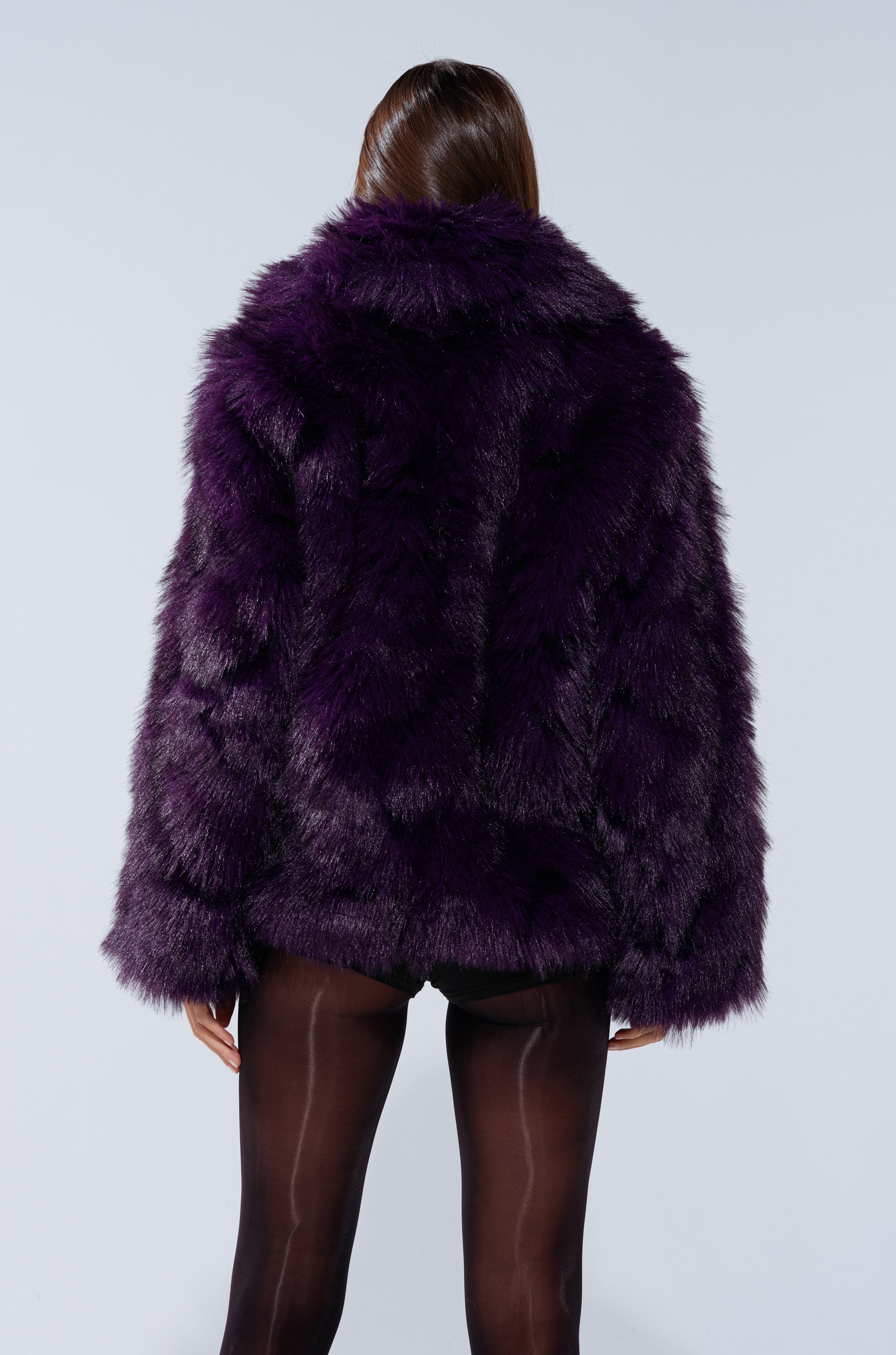 A person stands facing away, wearing the SHORTIE FUR COAT in oversized purple faux fur and sheer black tights against a plain light background.