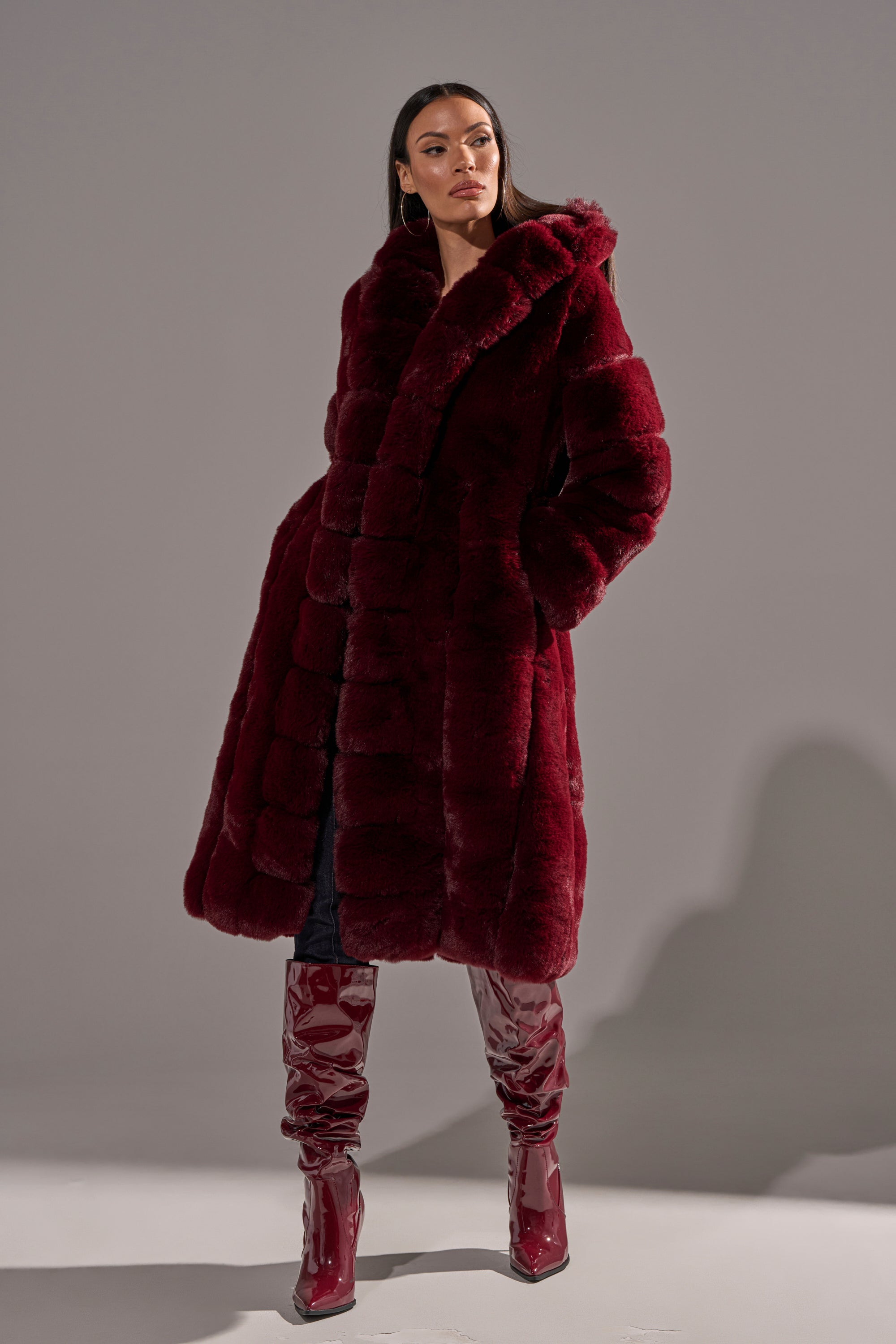 A woman with long dark hair poses confidently against a plain gray background, hands in her pockets, wearing the BERLIN HOODED FAUX FUR IN BURGUNDY coat styled with matching glossy burgundy knee-high boots.