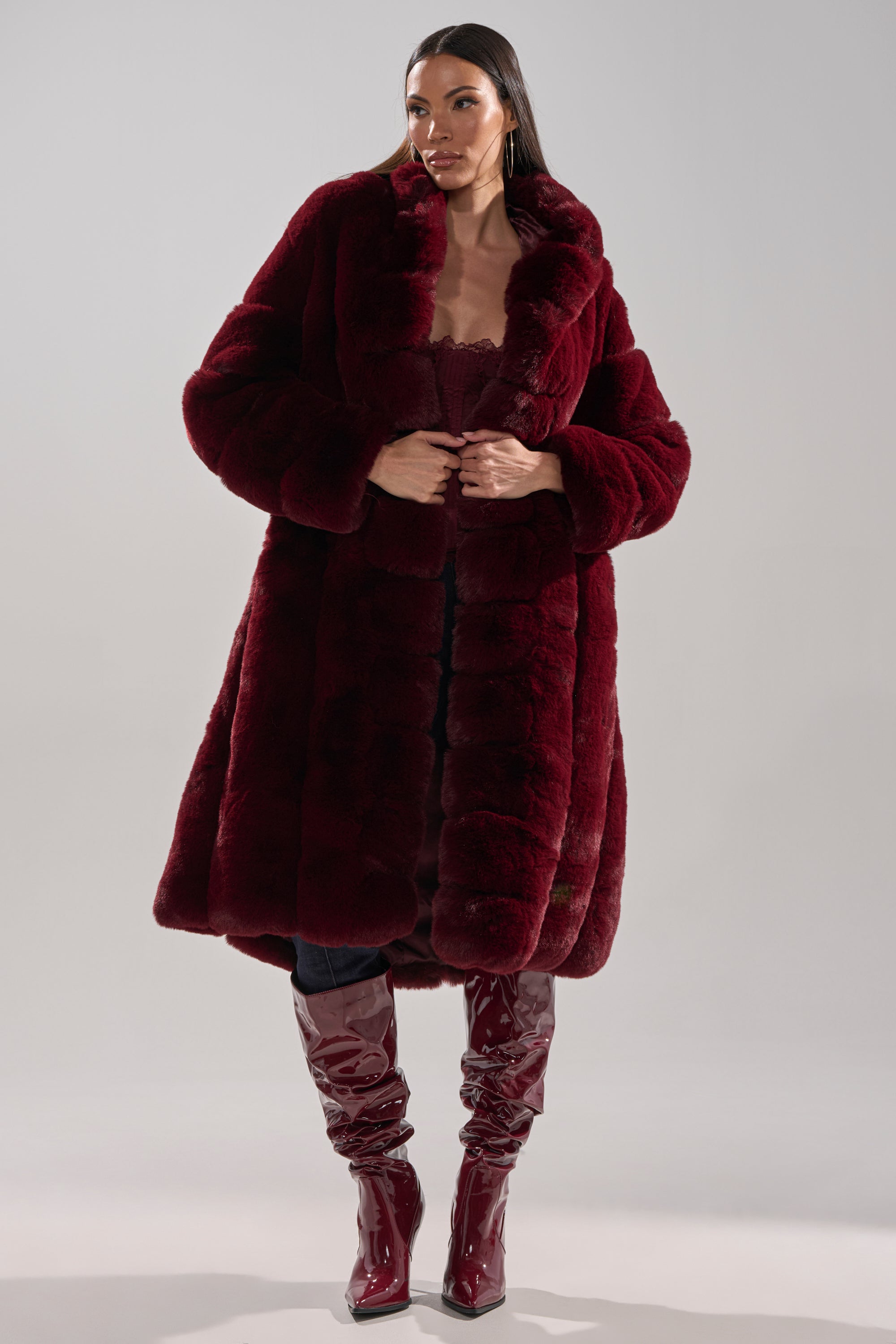 A woman wears the BERLIN HOODED FAUX FUR IN BURGUNDY, styled with dark pants, matching glossy knee-high boots, hoop earrings, and sleek straight hair, standing against a plain light background.