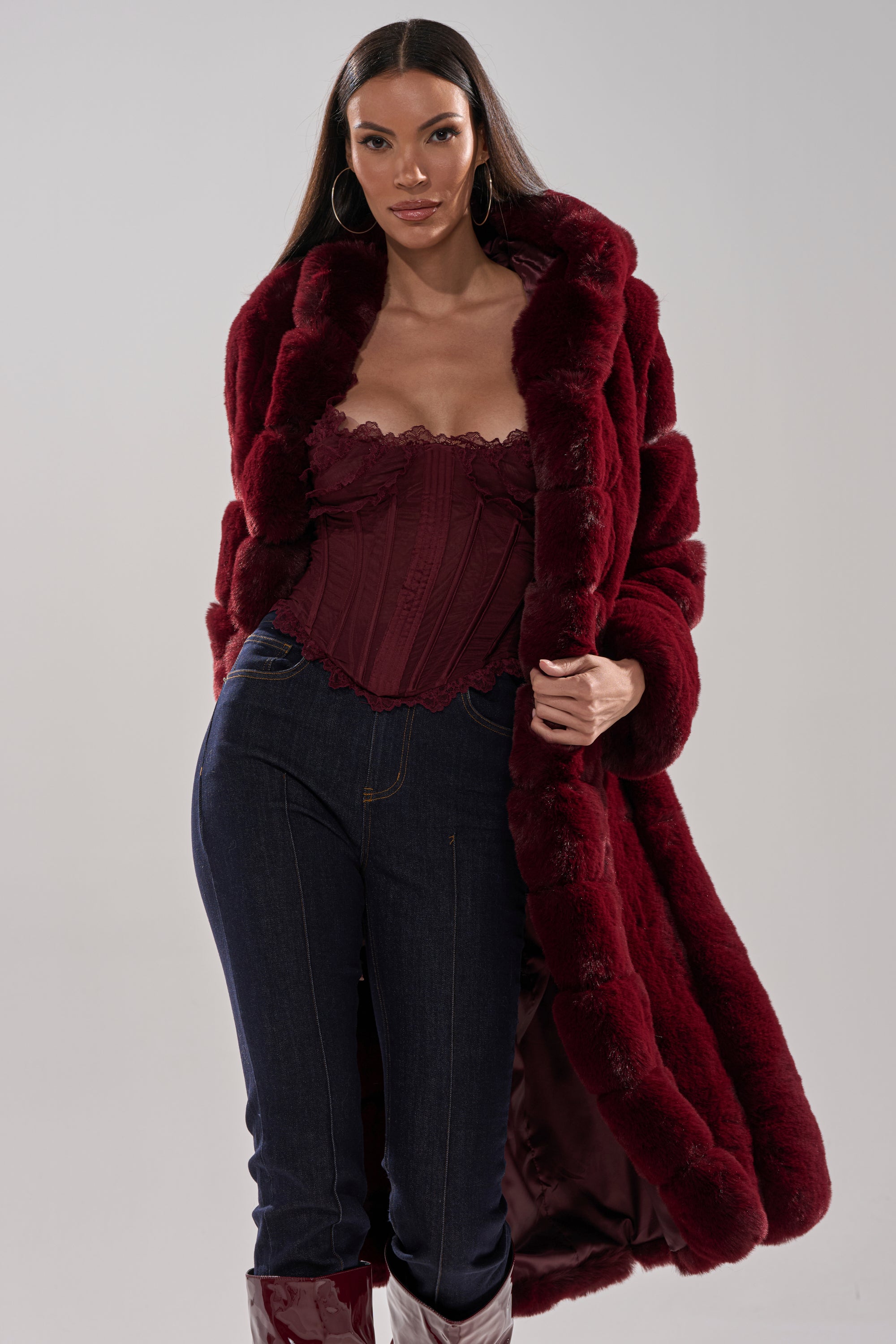 A woman stands confidently against a light gray background, wearing the BERLIN HOODED FAUX FUR IN BURGUNDY with a matching corset top, dark jeans, and red boots.