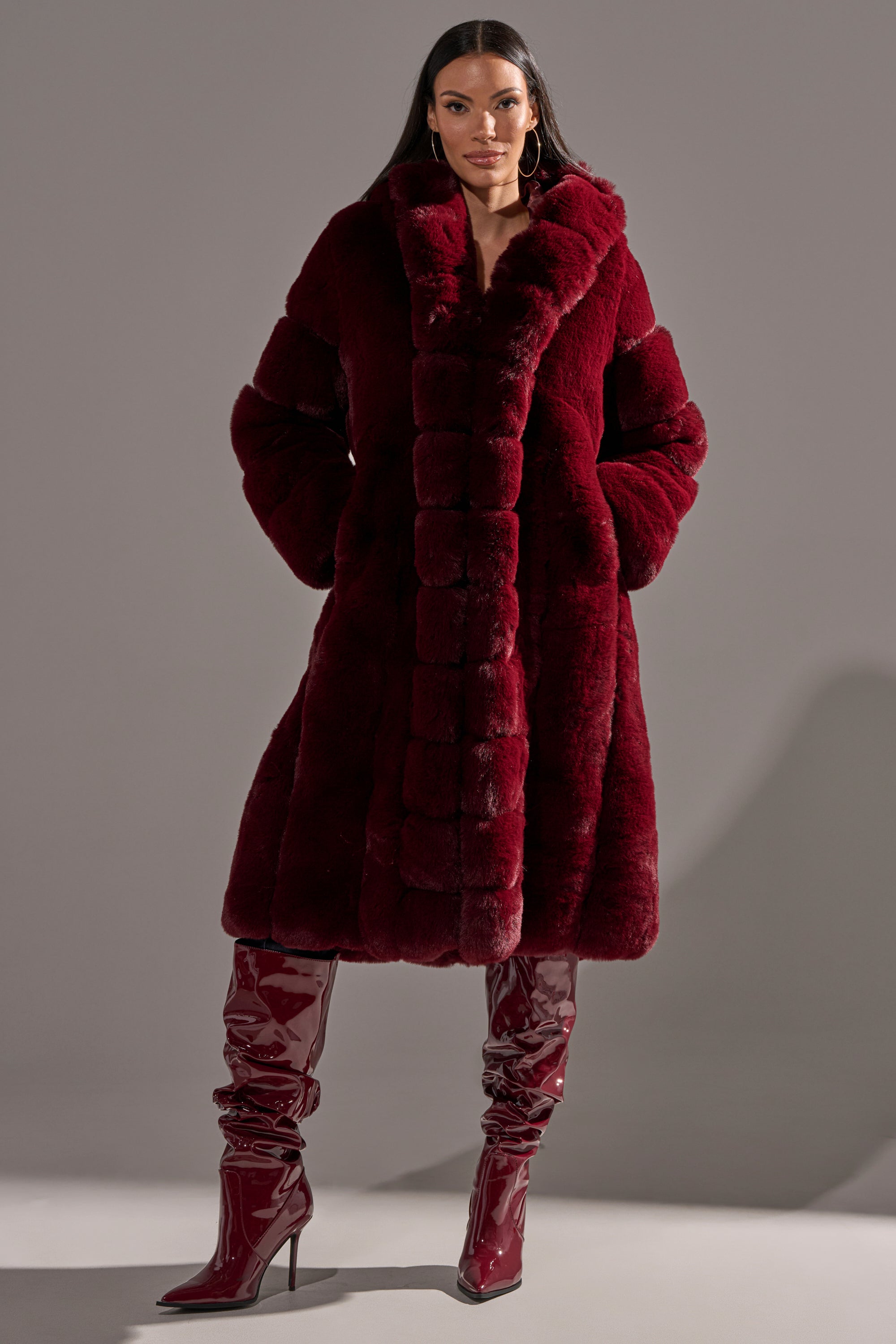 A woman stands confidently against a gray background, wearing the BERLIN HOODED FAUX FUR IN BURGUNDY and matching shiny red knee-high heeled boots.