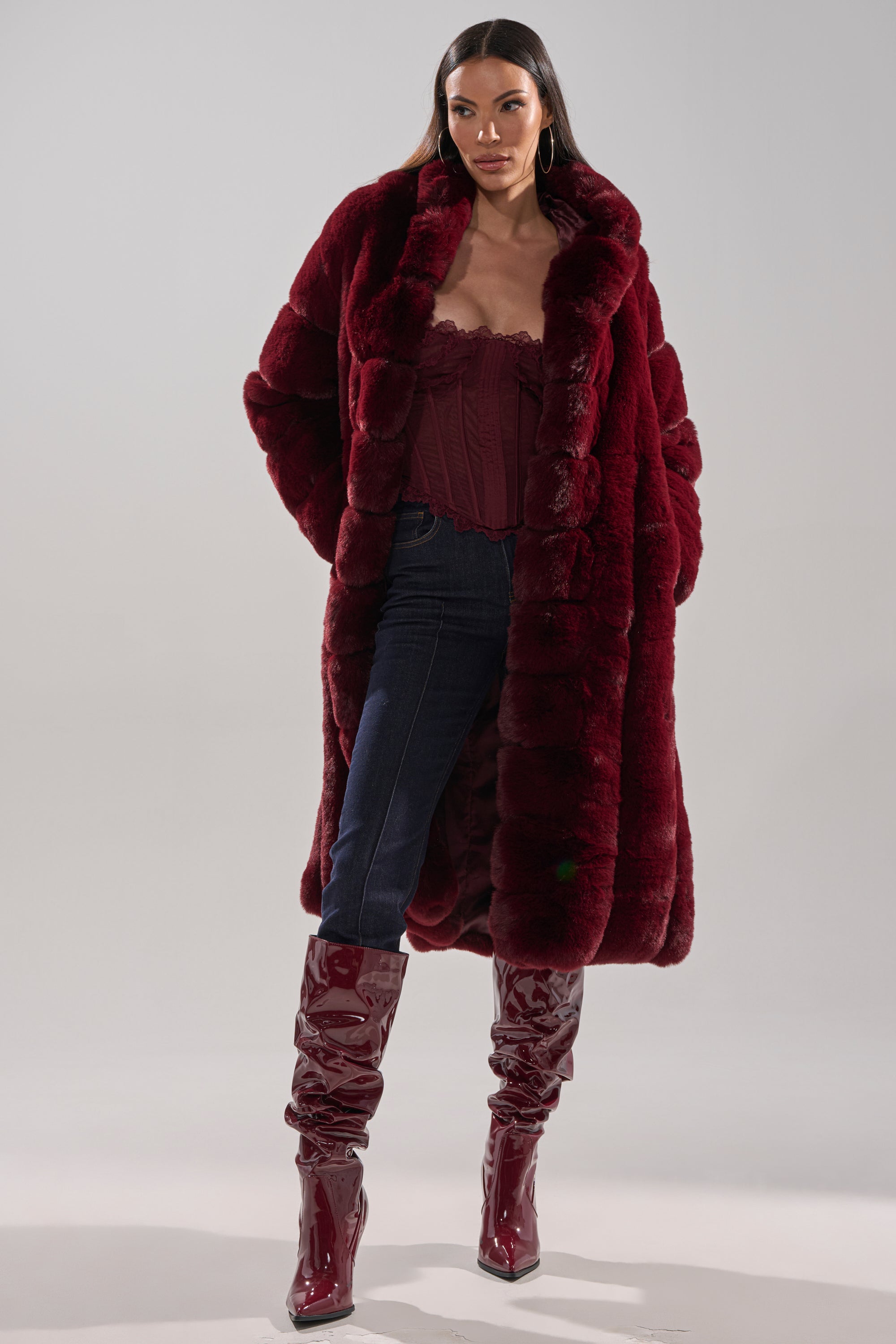 A woman stands confidently in the BERLIN HOODED FAUX FUR IN BURGUNDY, paired with a matching corset top, dark blue jeans, and shiny burgundy knee-high boots against a plain background.