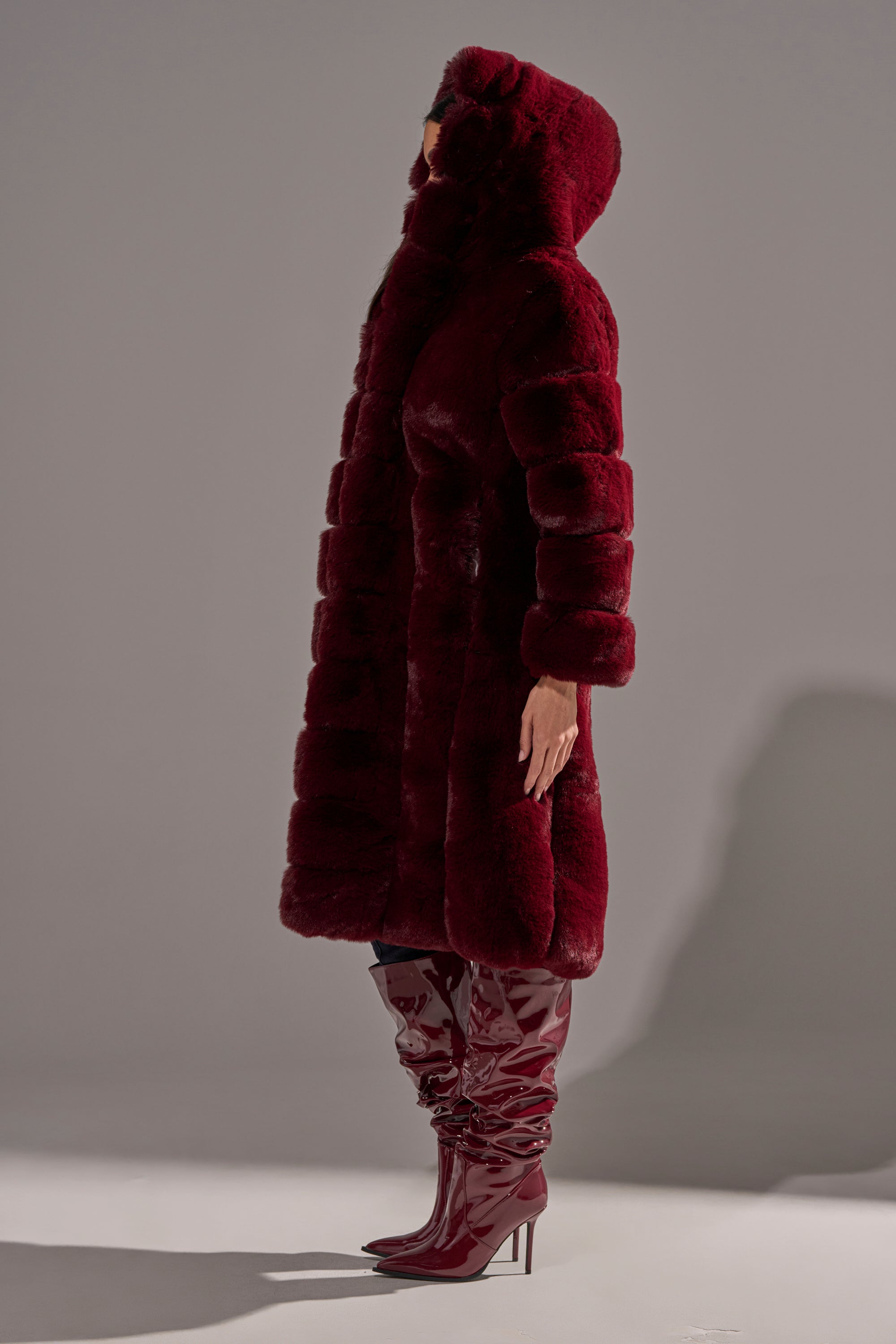 A person stands in profile against a plain gray background, wearing the BERLIN HOODED FAUX FUR IN BURGUNDY, a plush faux fur coat with a hood, paired with matching shiny burgundy knee-high, high-heeled boots.