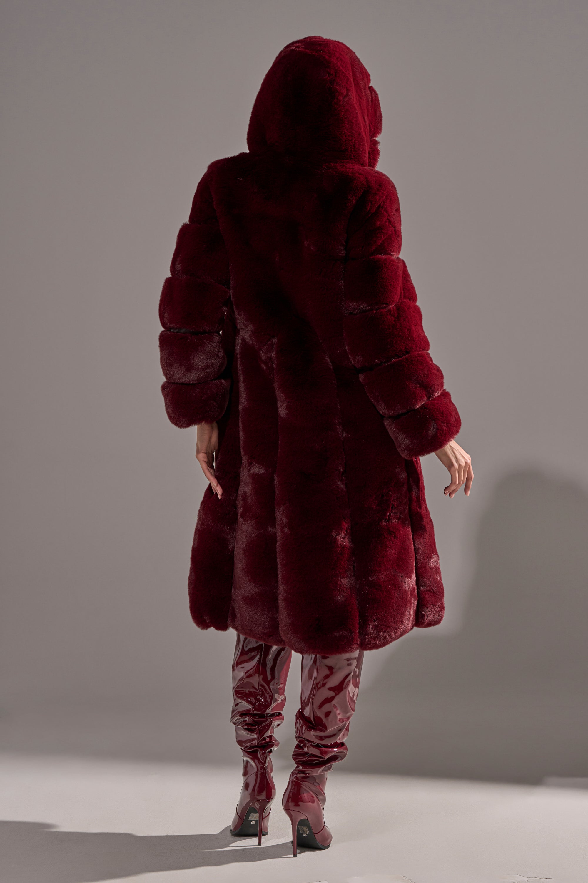 Wearing the BERLIN HOODED FAUX FUR IN BURGUNDY, a person stands with their back to the camera; the plush hooded coat’s rich color contrasts dramatically with the neutral background and pairs with shiny red high-heeled boots.