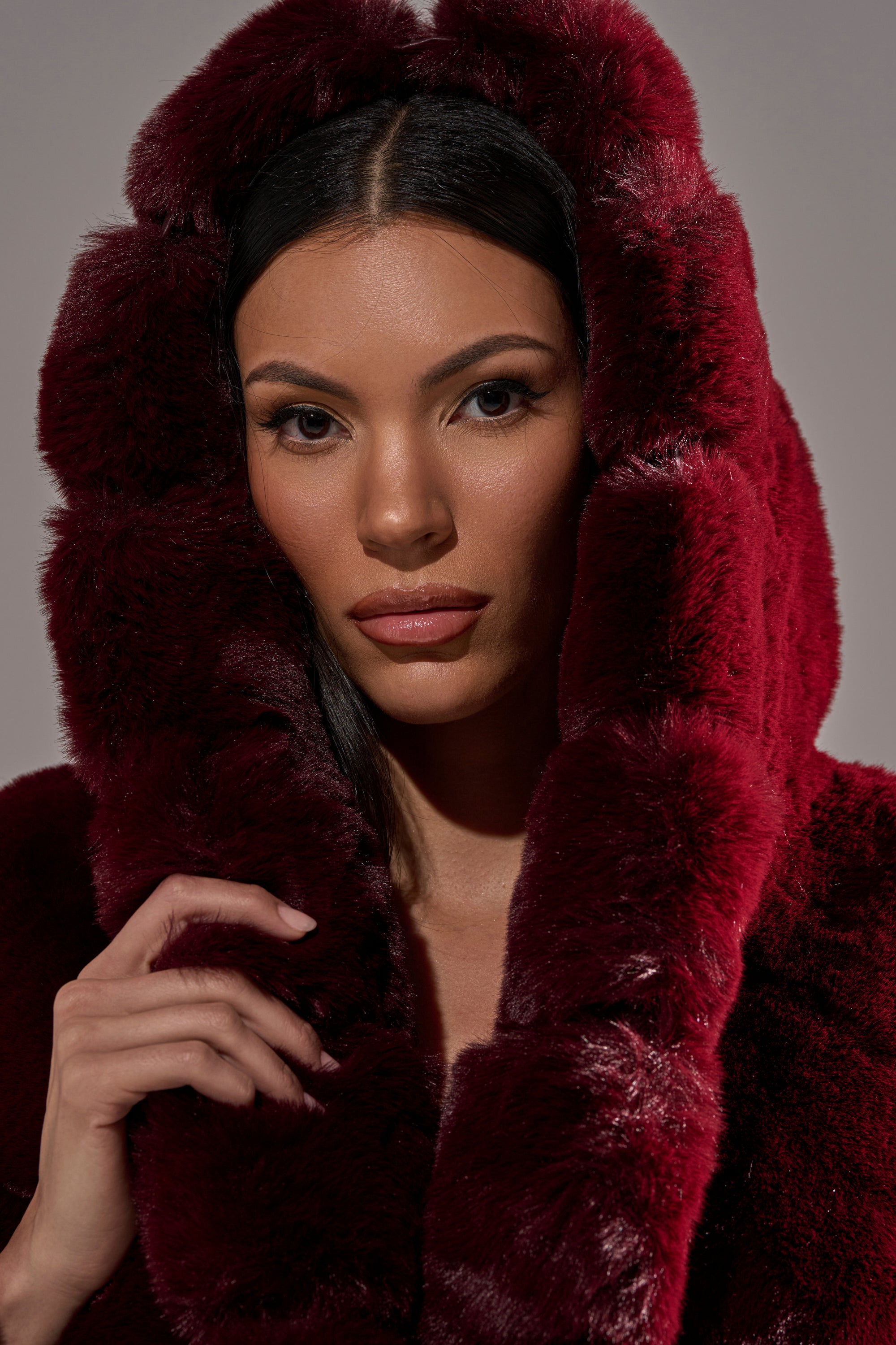 A woman with long dark hair wears the BERLIN HOODED FAUX FUR IN BURGUNDY, gazing at the camera with a neutral expression while holding the hood near her face.